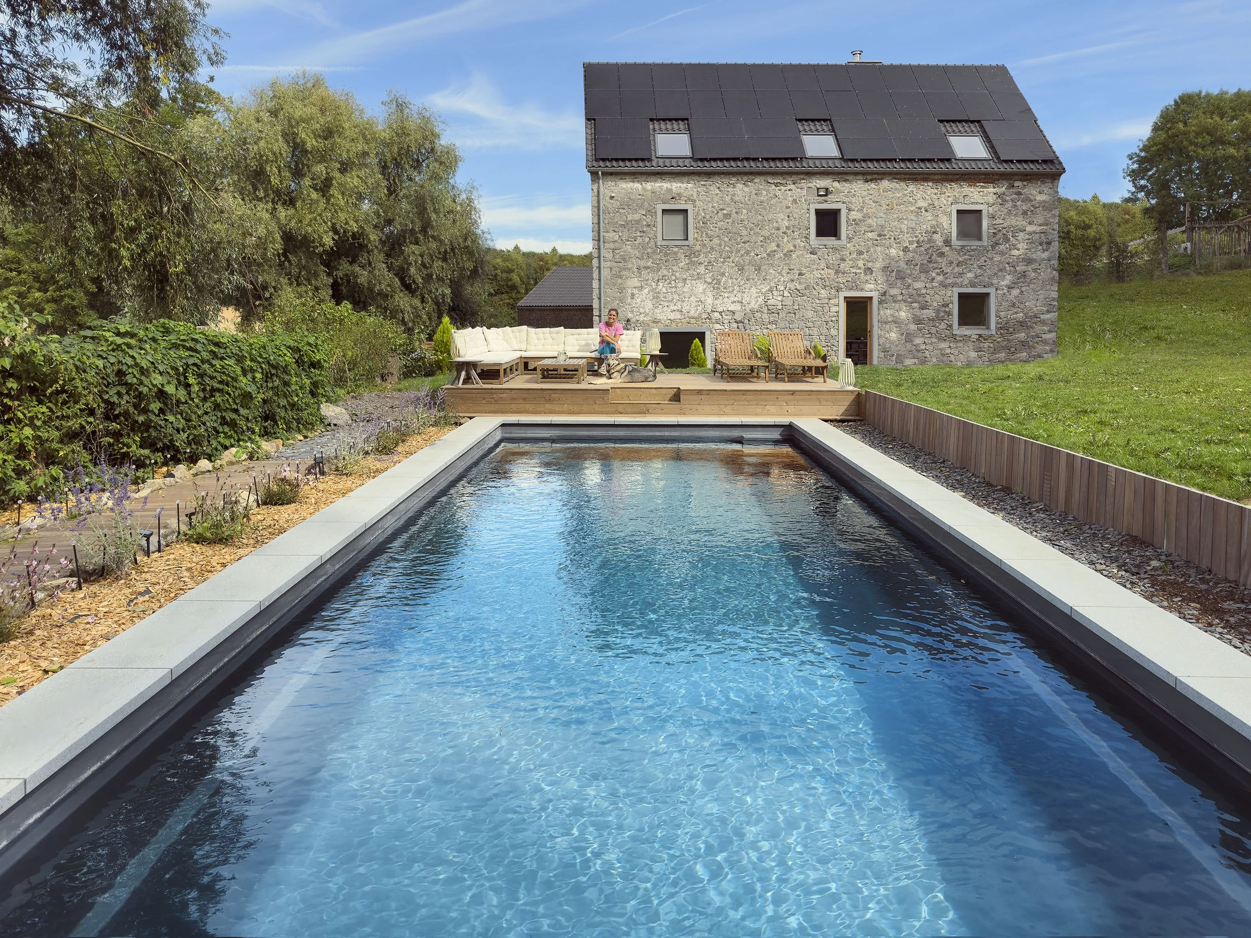 Piscine rectangulaire dans un jardin, avec un massif de plantes décoratives sur le côté gauche et une maison en pierre à l'arrière-plan, sous un ciel bleu avec quelques nuages.
