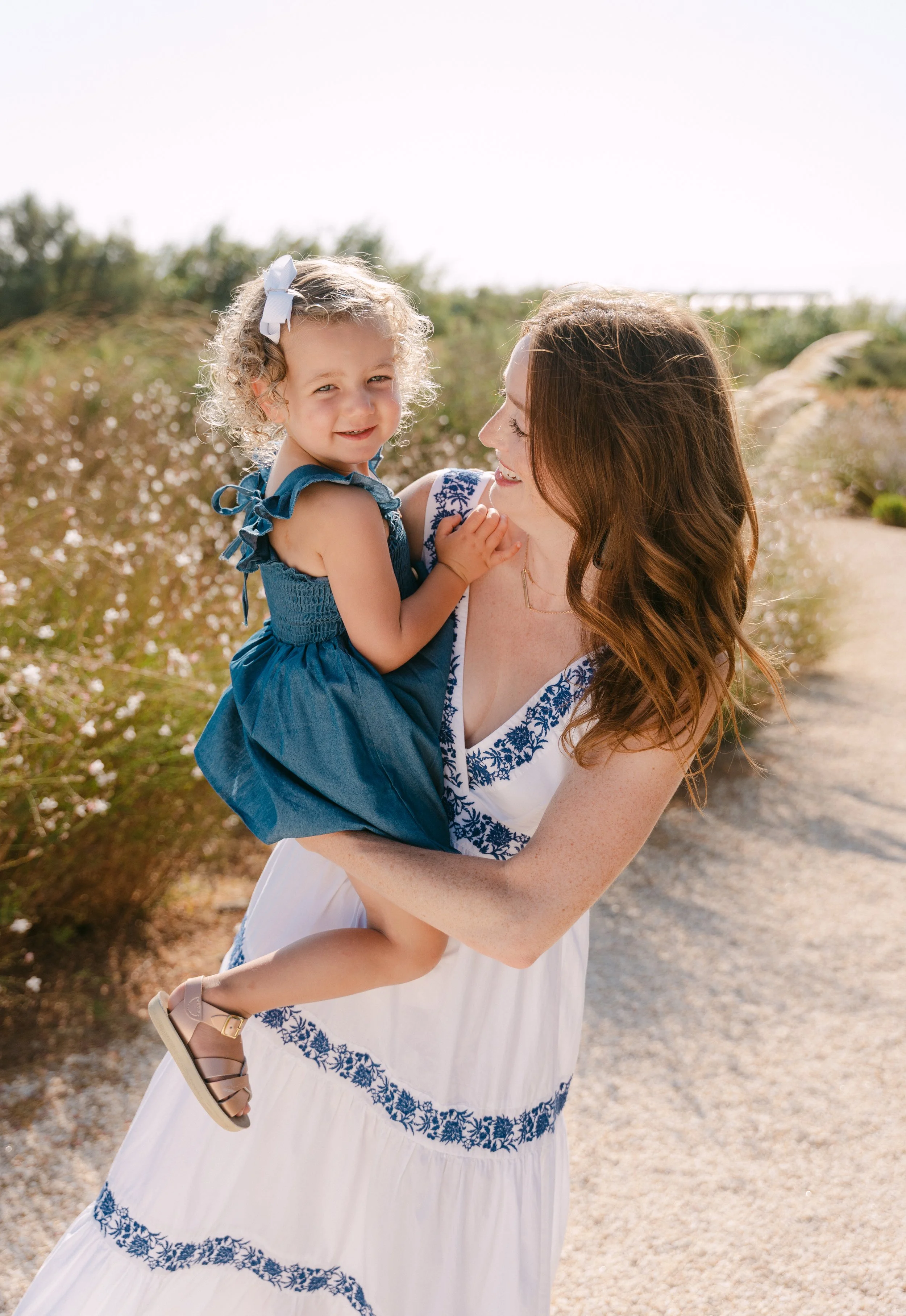 A woman with red hair and a white dress with blue embroidery is holding a young girl with curly blonde hair and a blue dress outdoors on a sunny day, with flowering bushes and a gravel path in the background.