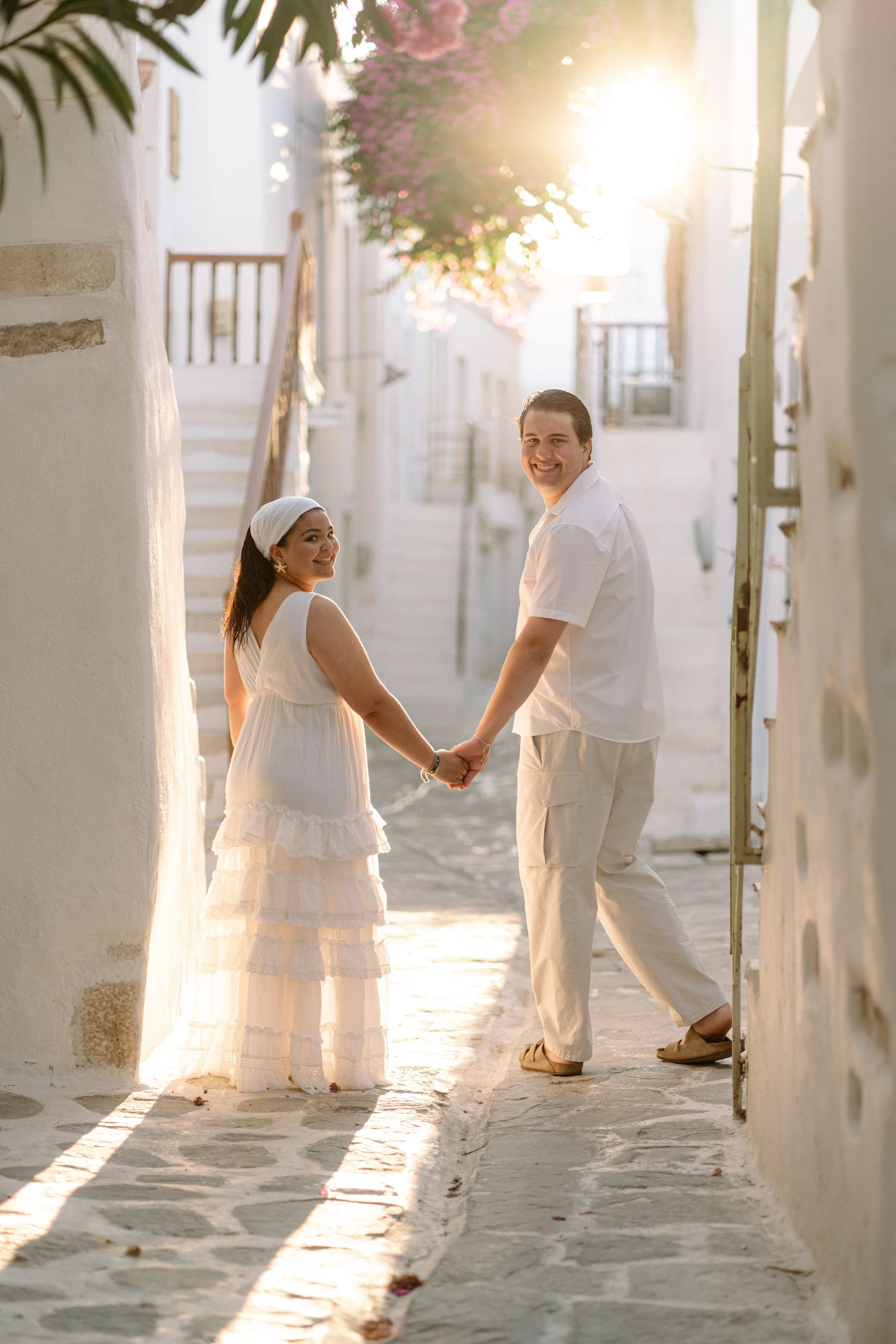 A happy couple holding hands and smiling in an alleyway with white buildings, purple flowers, and warm sunlight.
