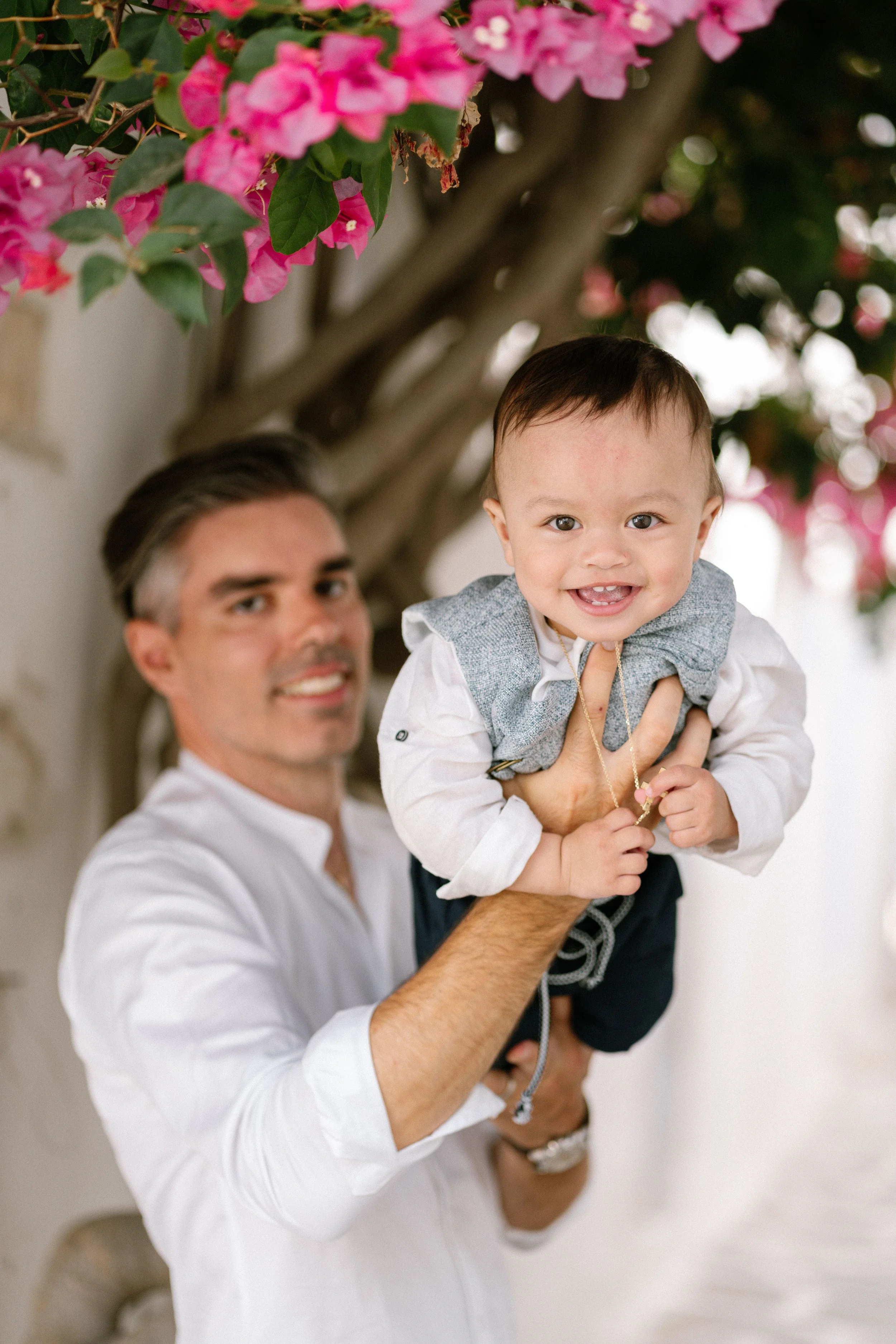 A smiling toddler holding a man outdoors beneath a pink flowering tree.
