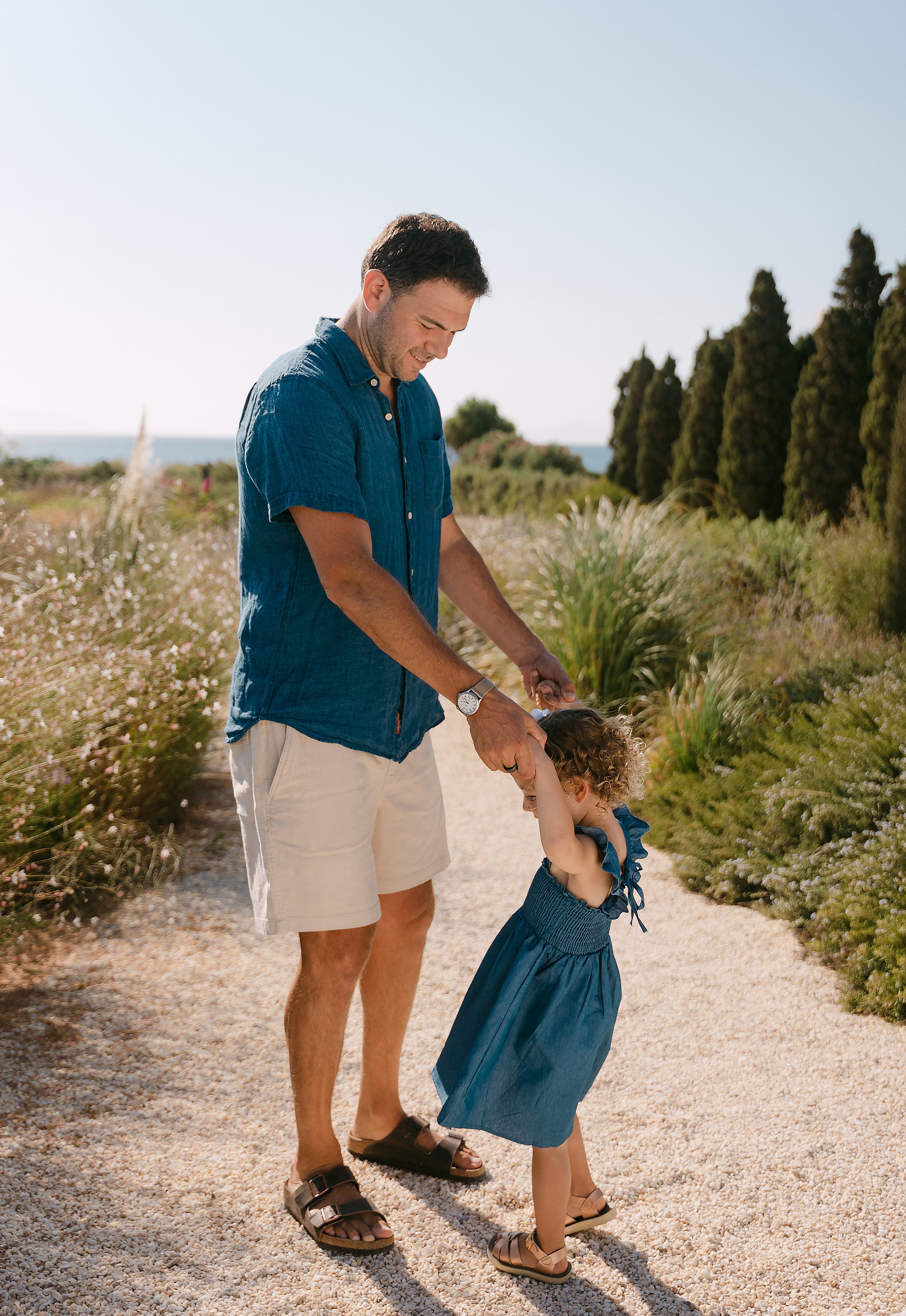 A man with short dark hair, wearing a blue short-sleeve shirt and beige shorts, is holding a young girl’s hands as she twirls and laughs on a sunny path near the coast. The girl has curly hair and is wearing a blue dress and sandals, with tall bushes