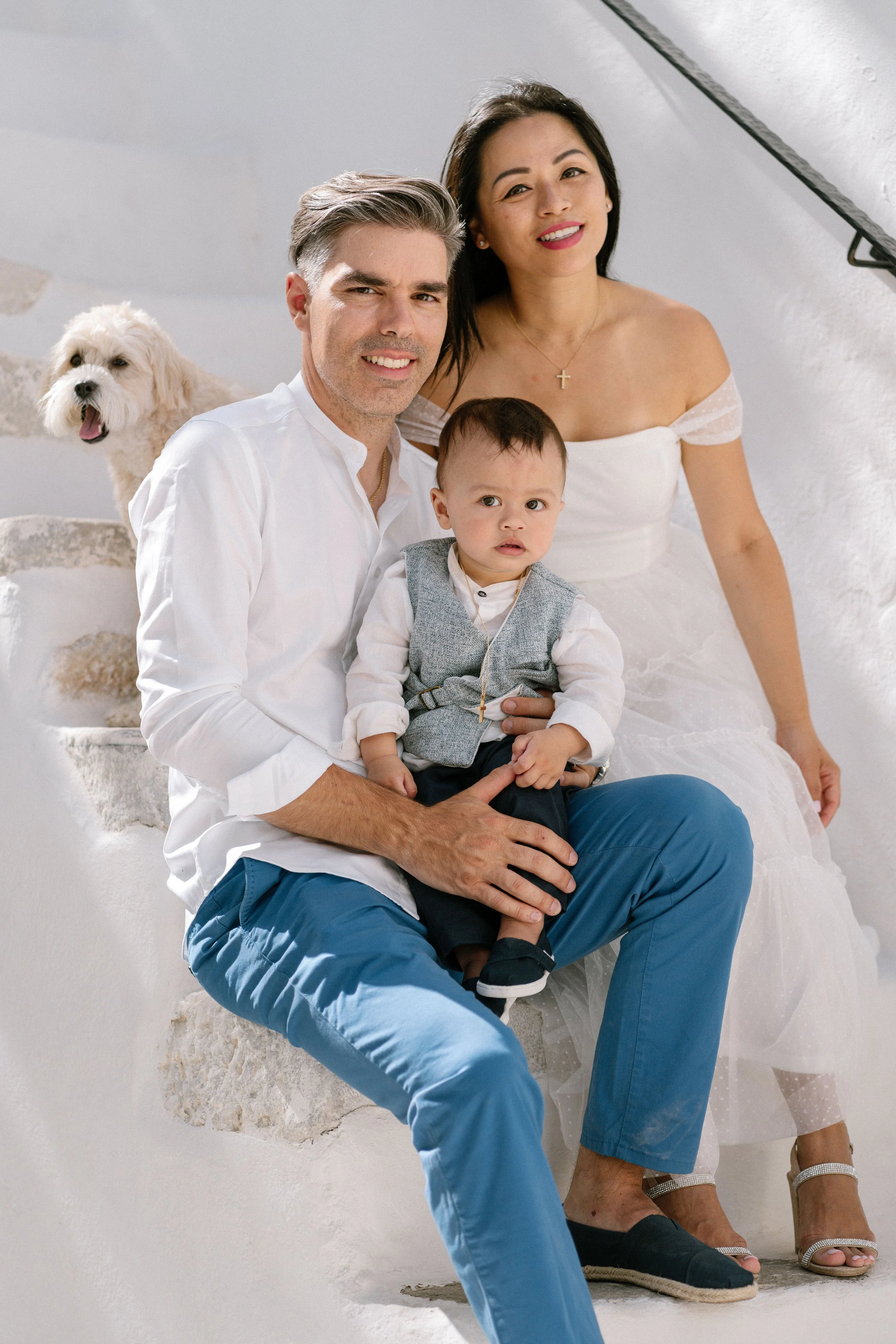 A family of three sitting on white stone stairs with a small dog in the background. The man is holding a young boy. The woman is sitting behind them. The family is dressed in semi-formal attire, with the man and boy wearing white shirts, and the woma