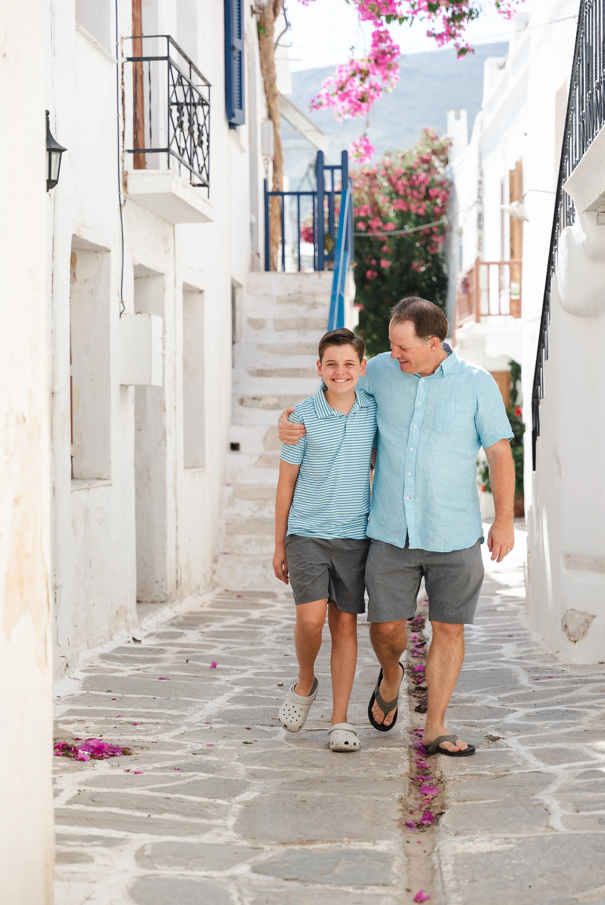 A smiling boy and man walk arm-in-arm down a whitewashed alleyway with stone pavement, pink bougainvillea flowers on trees, and Greek-style architecture.