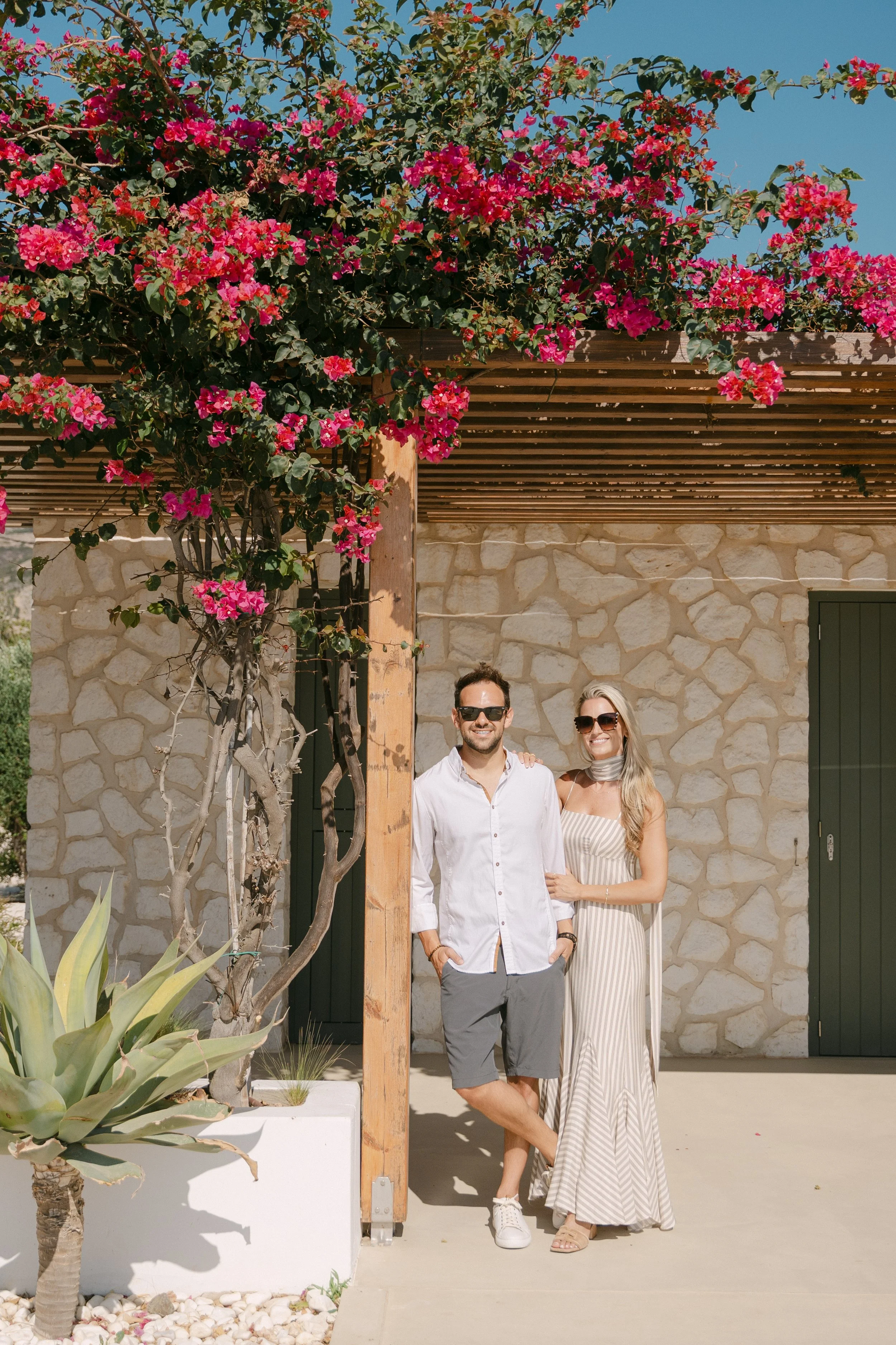 A man and woman stand close together outdoors in front of a stone wall; the man is wearing a white shirt and shorts, the woman is in a cream-colored striped dress. They are smiling and wearing sunglasses, with pink flowering vines above them.