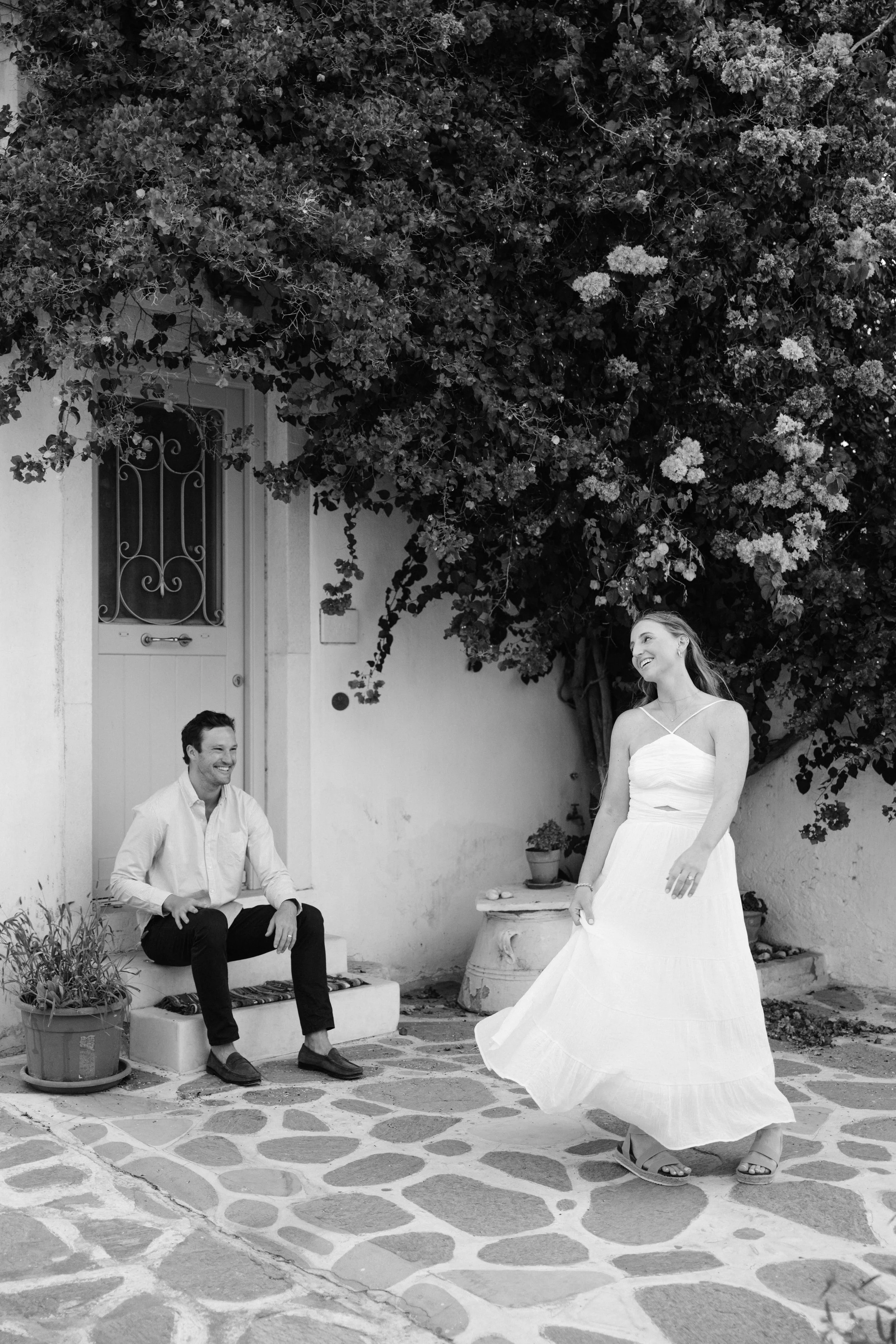 A woman in a white dress dancing happily while a man sits on a step smiling at her, outside a house with potted plants and a tree with dense foliage.