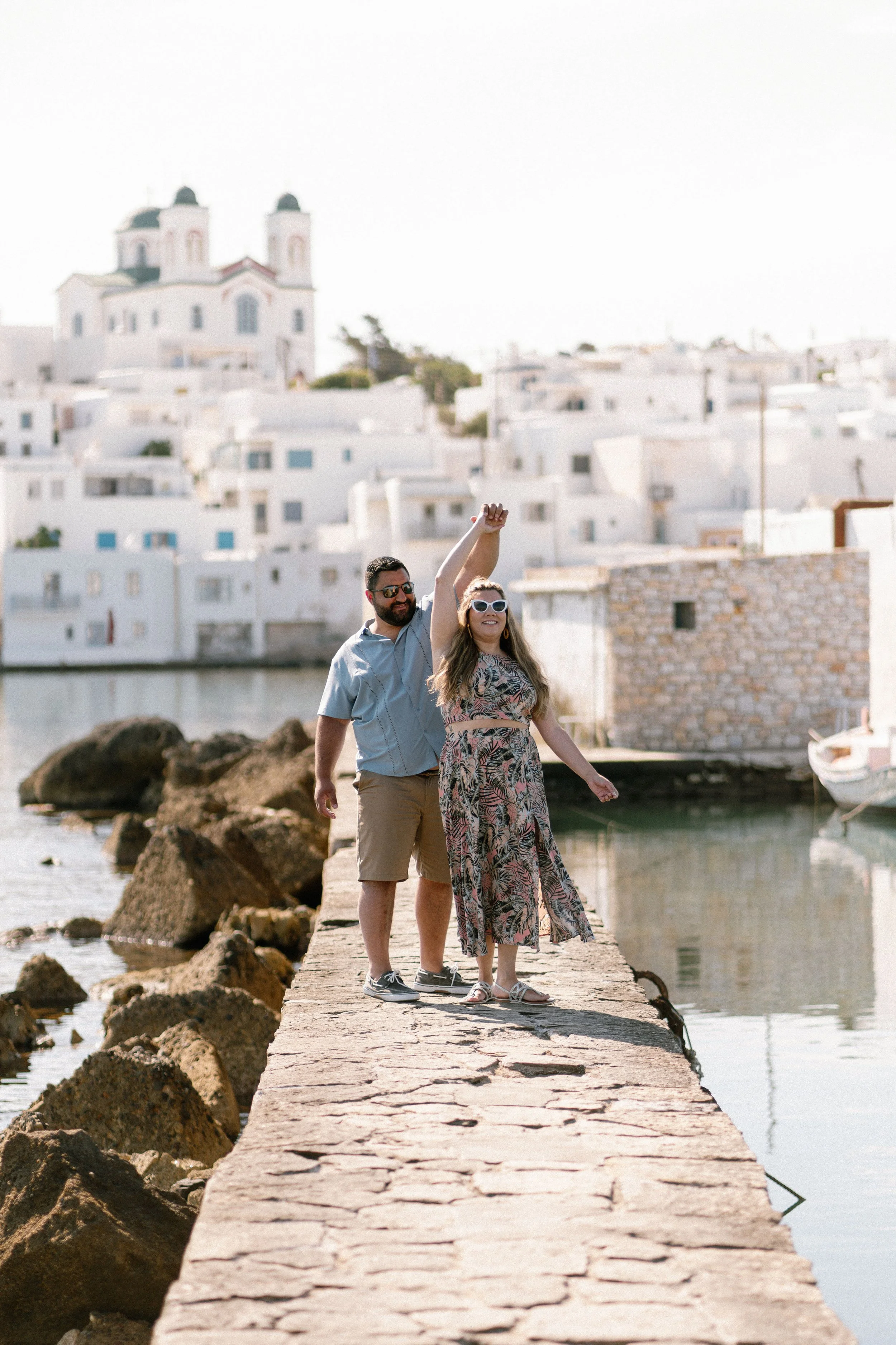 A man and woman standing on a stone pier by the water, with white buildings and a church in the background. The woman is raising her arm, and both are smiling and wearing sunglasses.