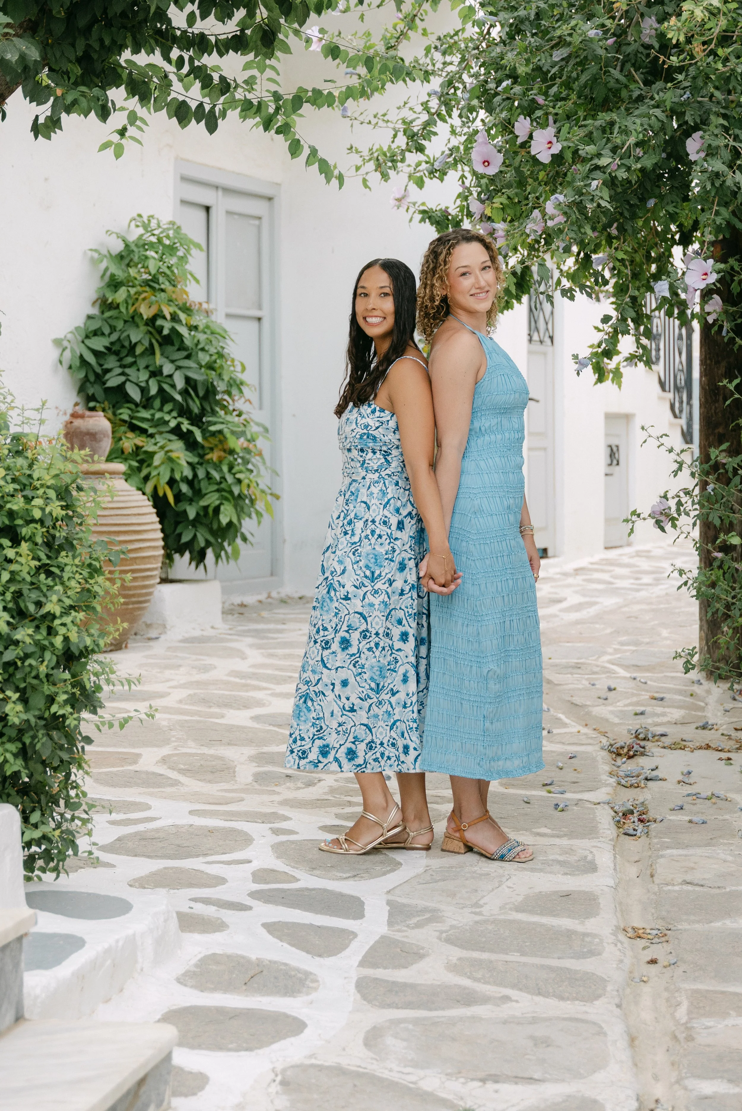Two women in blue dresses holding hands, standing back to back outdoors with greenery and white buildings in the background.