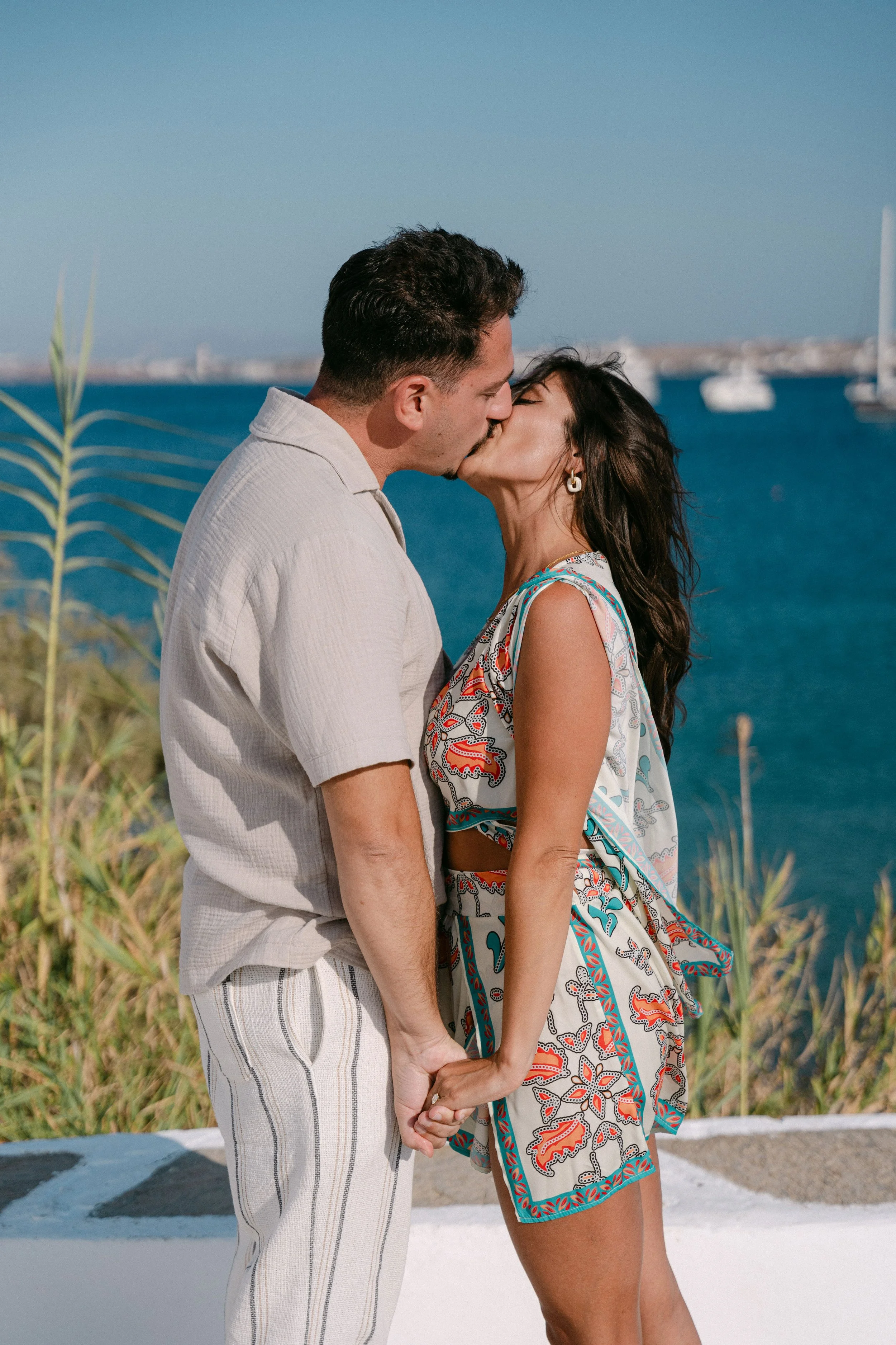 Couple kissing by the sea, holding hands, with boats in the background.