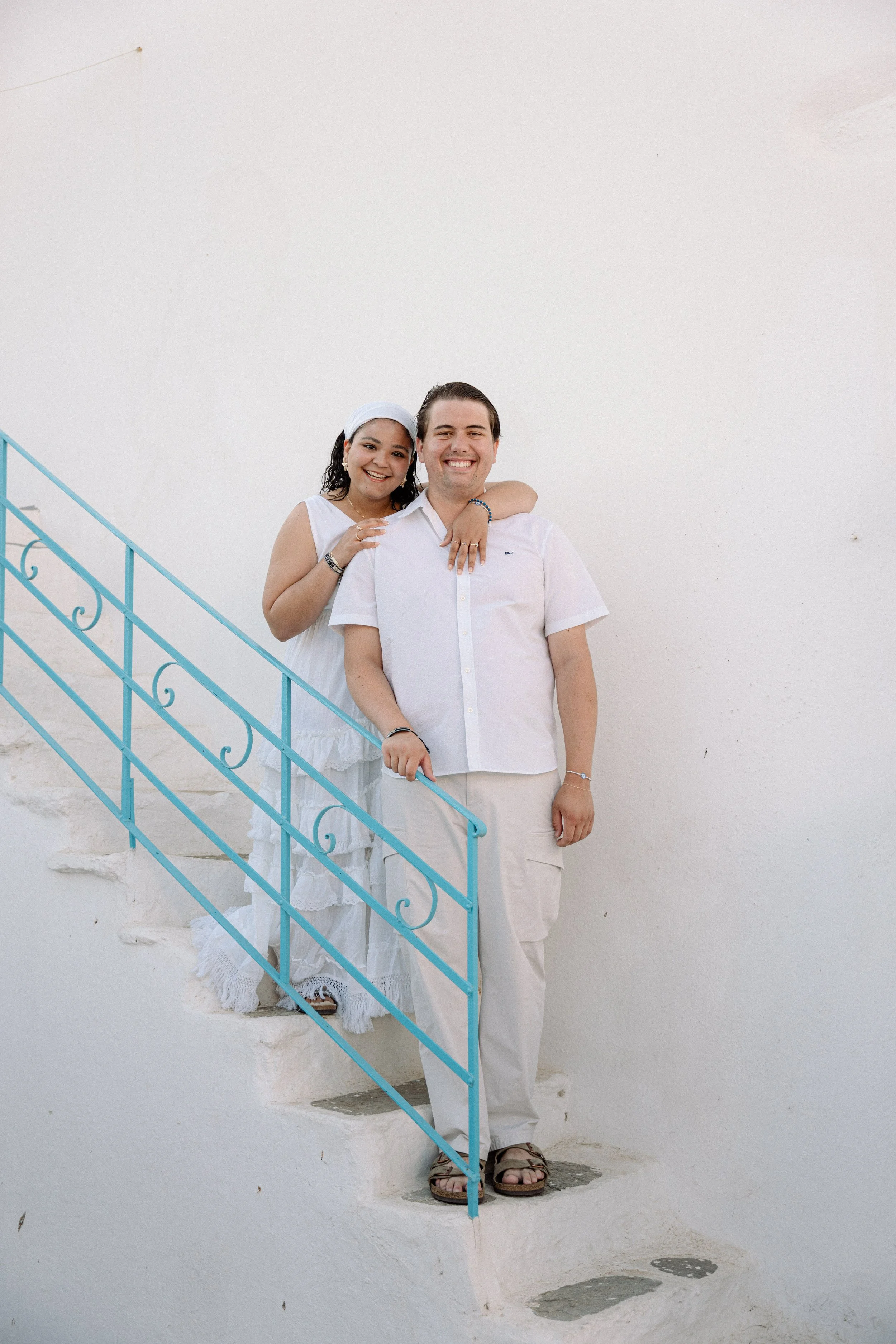 A smiling couple standing on white stairs with a turquoise railing against a white wall, dressed in white casual clothing.