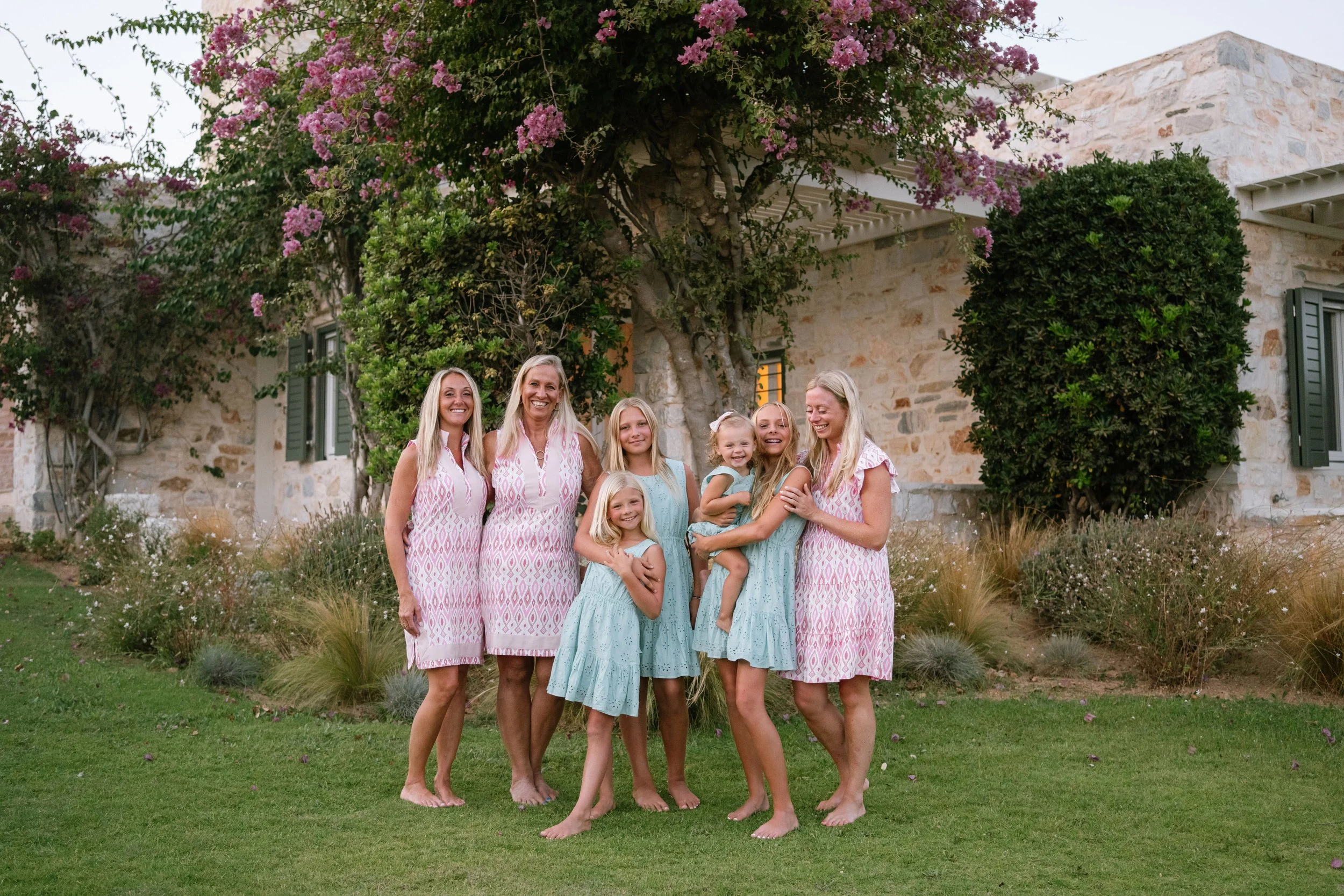 A group of seven women and girls standing on a lawn in front of a stone house with purple flowering trees and bushes, smiling and enjoying a moment together.