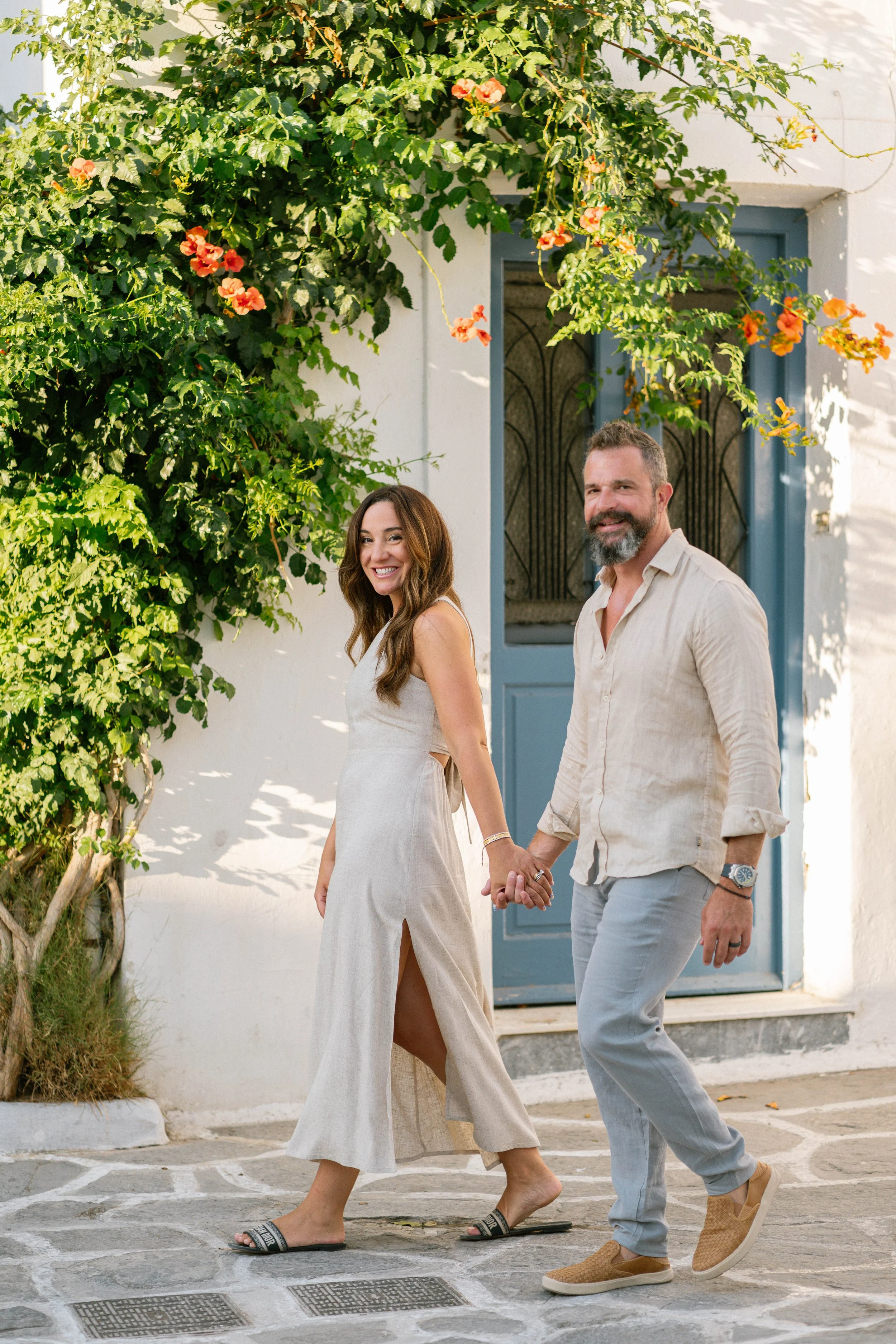 A smiling couple holding hands walking on a cobblestone street near a white building with a blue door and orange flowers climbing on a vine.