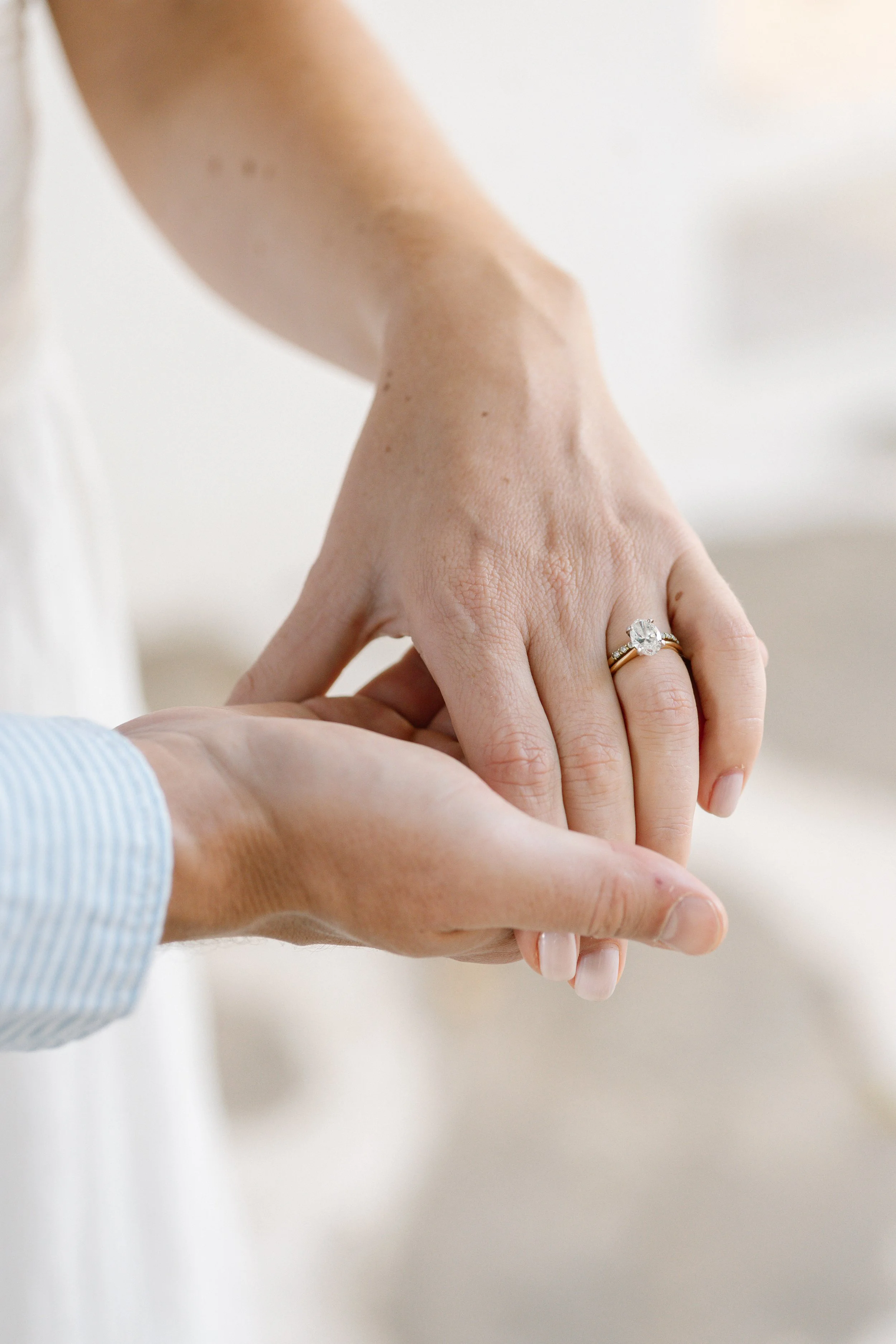 Close-up of a woman’s hand wearing an engagement ring, holding a man’s hand with a blue striped shirt. The background is blurred and light-colored.