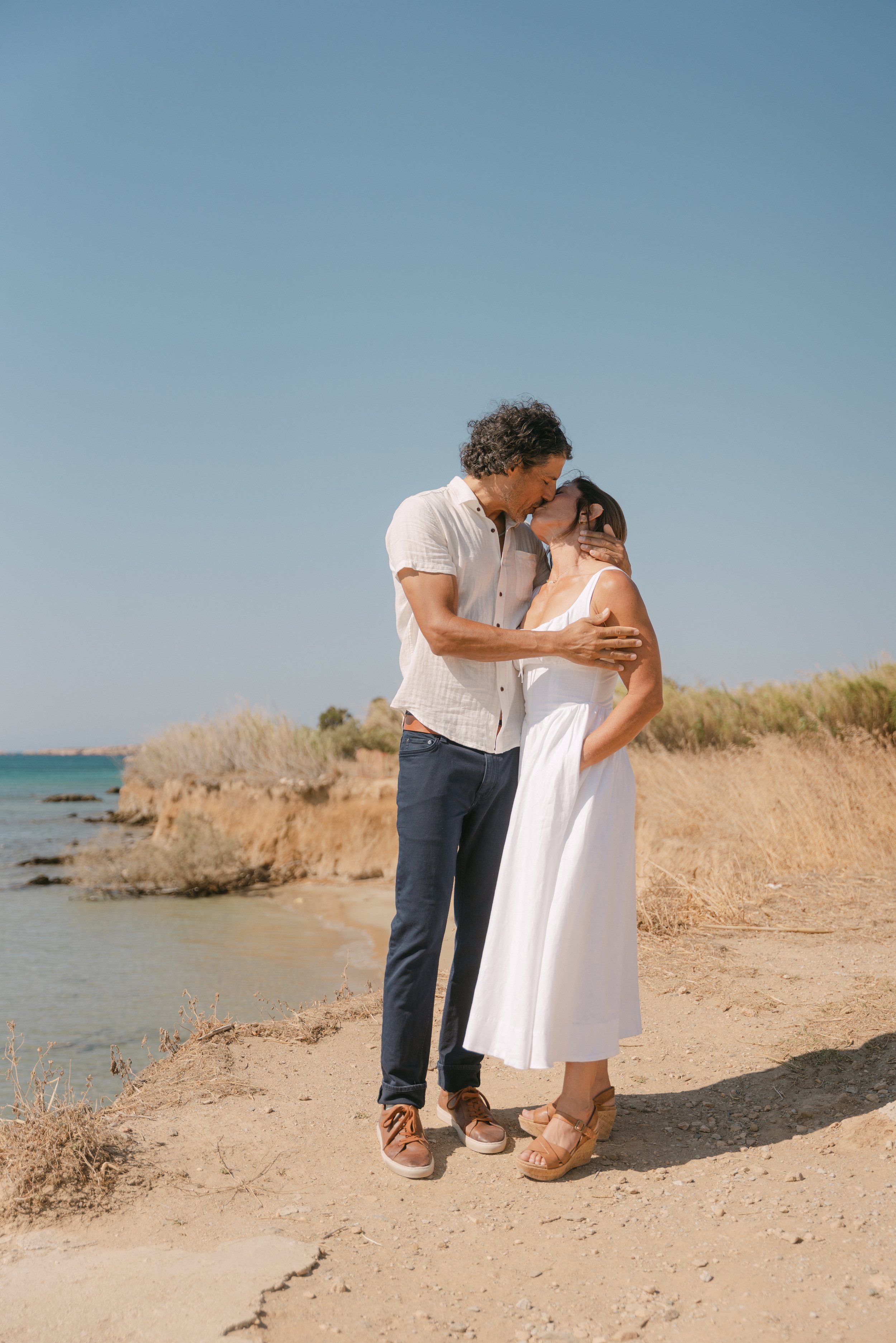 A couple kissing on a beach, with the man holding the woman's face and the woman with her hand on her hip, under a clear blue sky.