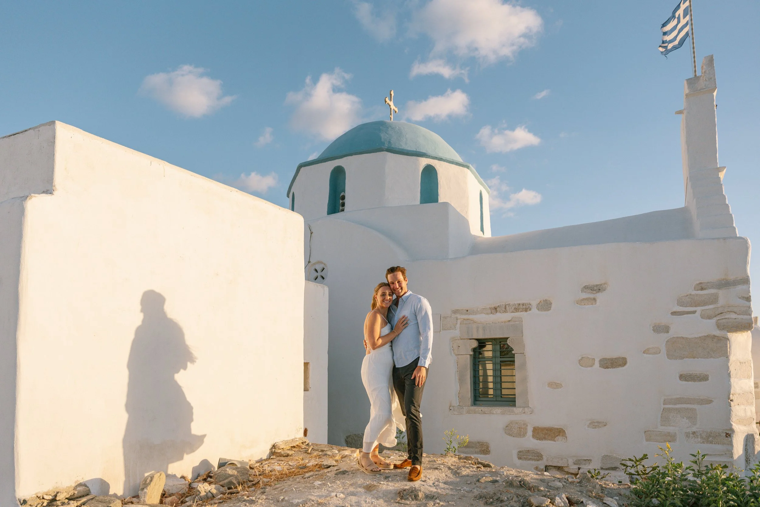 A couple standing close together outside a white chapel with a blue dome, a Greek flag flying, and a cross on top, in Greece during sunset.