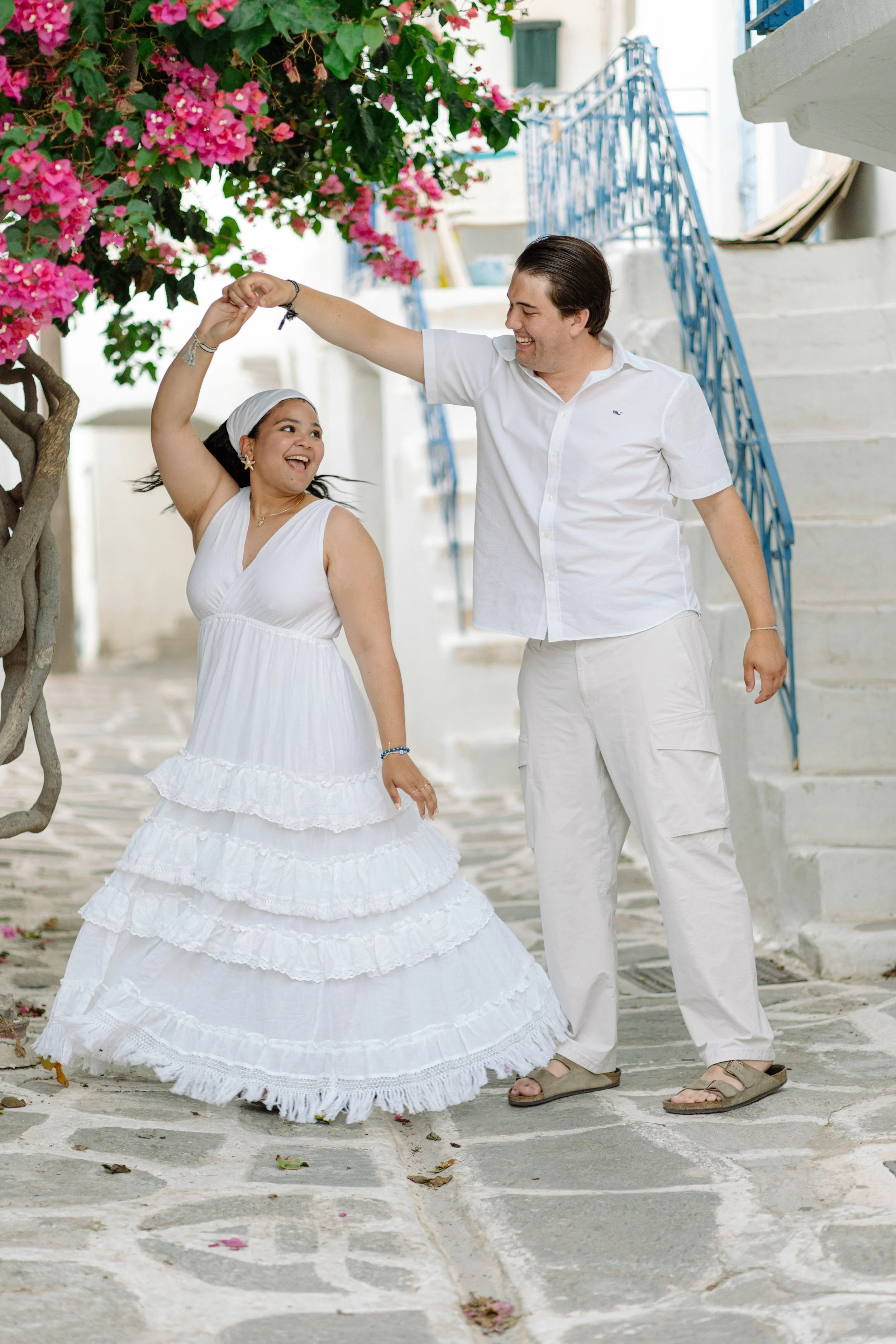 A smiling woman in a white, tiered, summer dress dancing with a man in a white shirt and beige pants under a pink flowering tree on a stone-paved street, with white buildings and blue railings in the background.