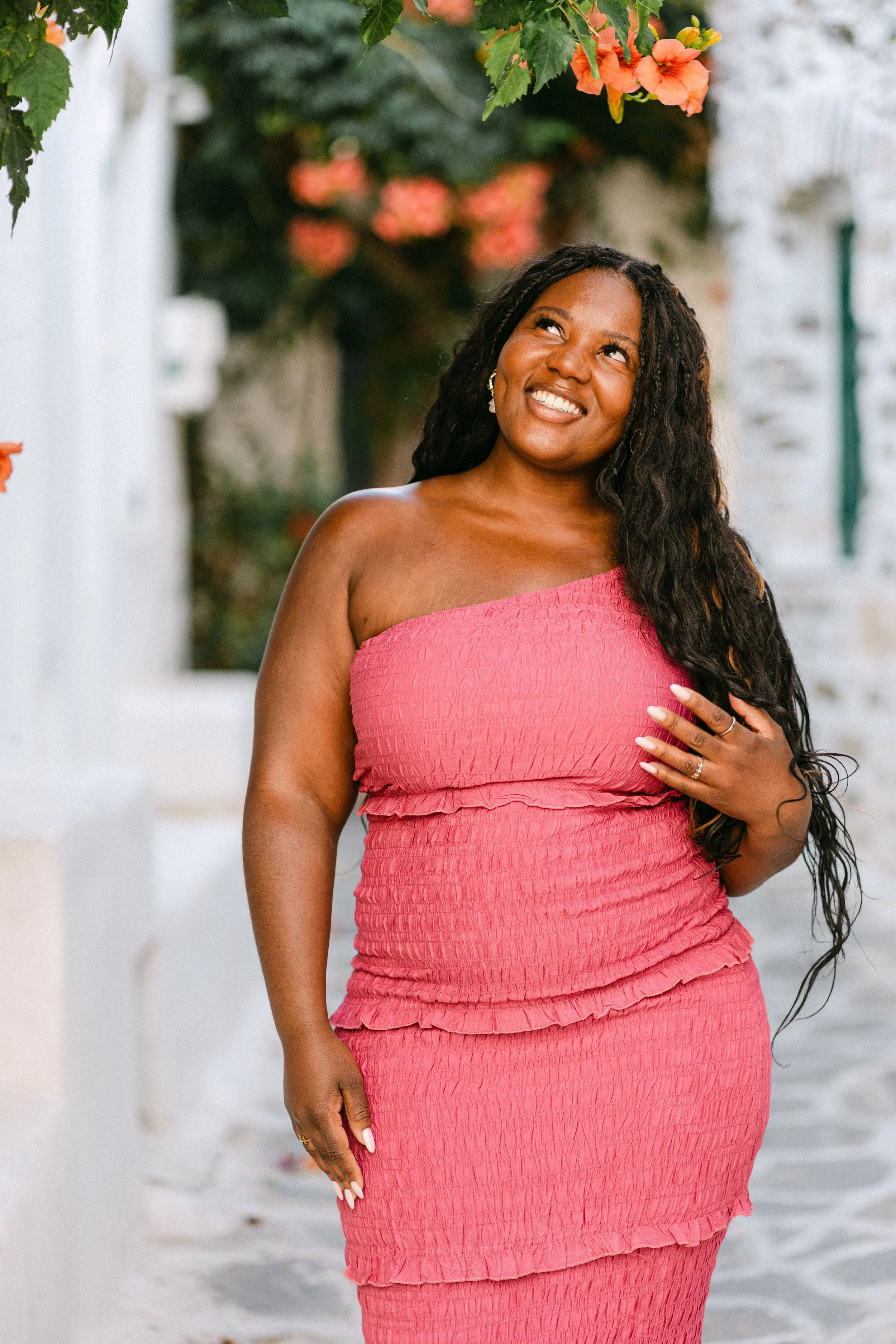 A woman with long curly hair in a pink strapless dress standing outdoors in a white alleyway with flowers and greenery, smiling and looking up.