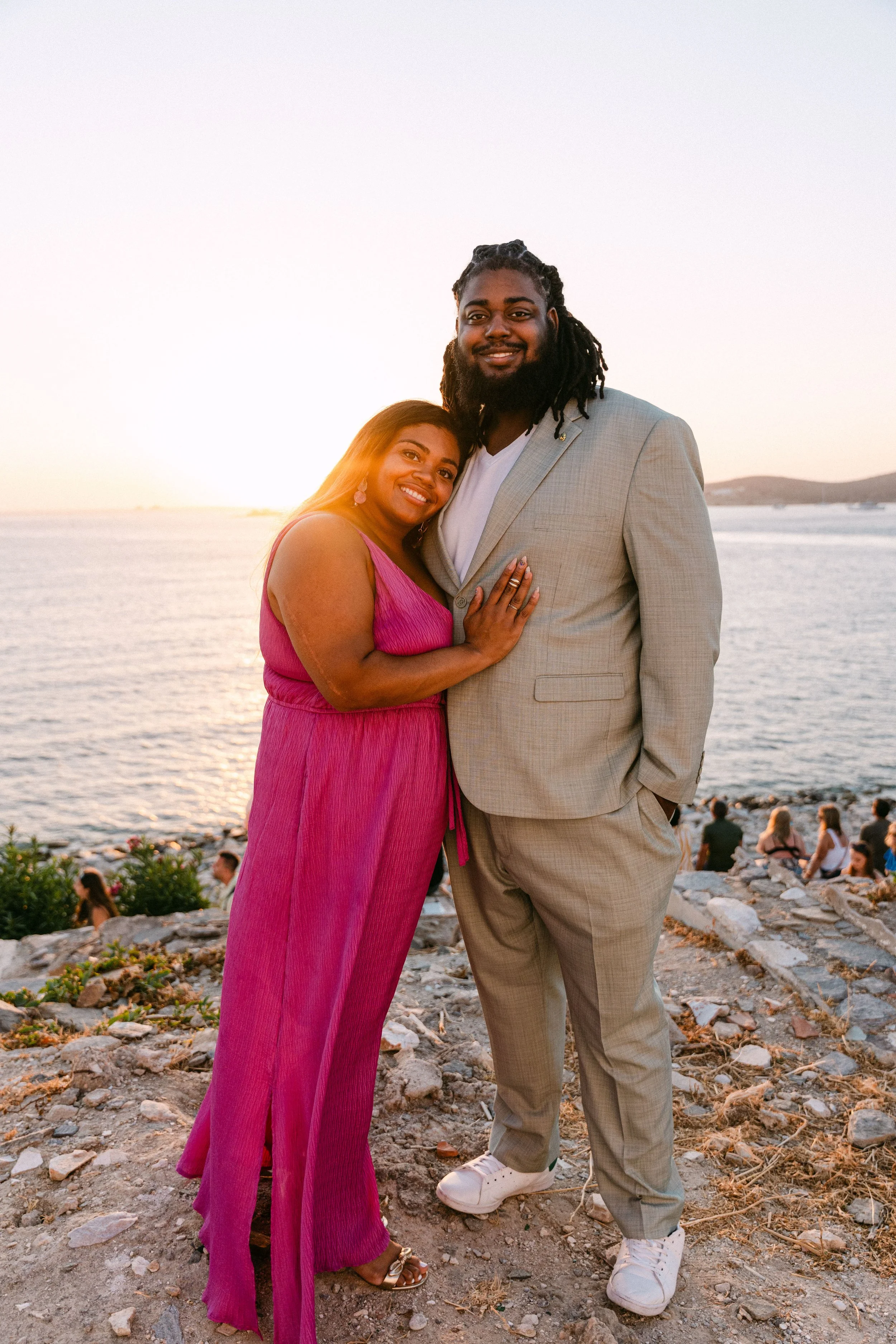 A smiling couple standing on a rocky area near water at sunset, the woman in a pink dress and the man in a beige suit.