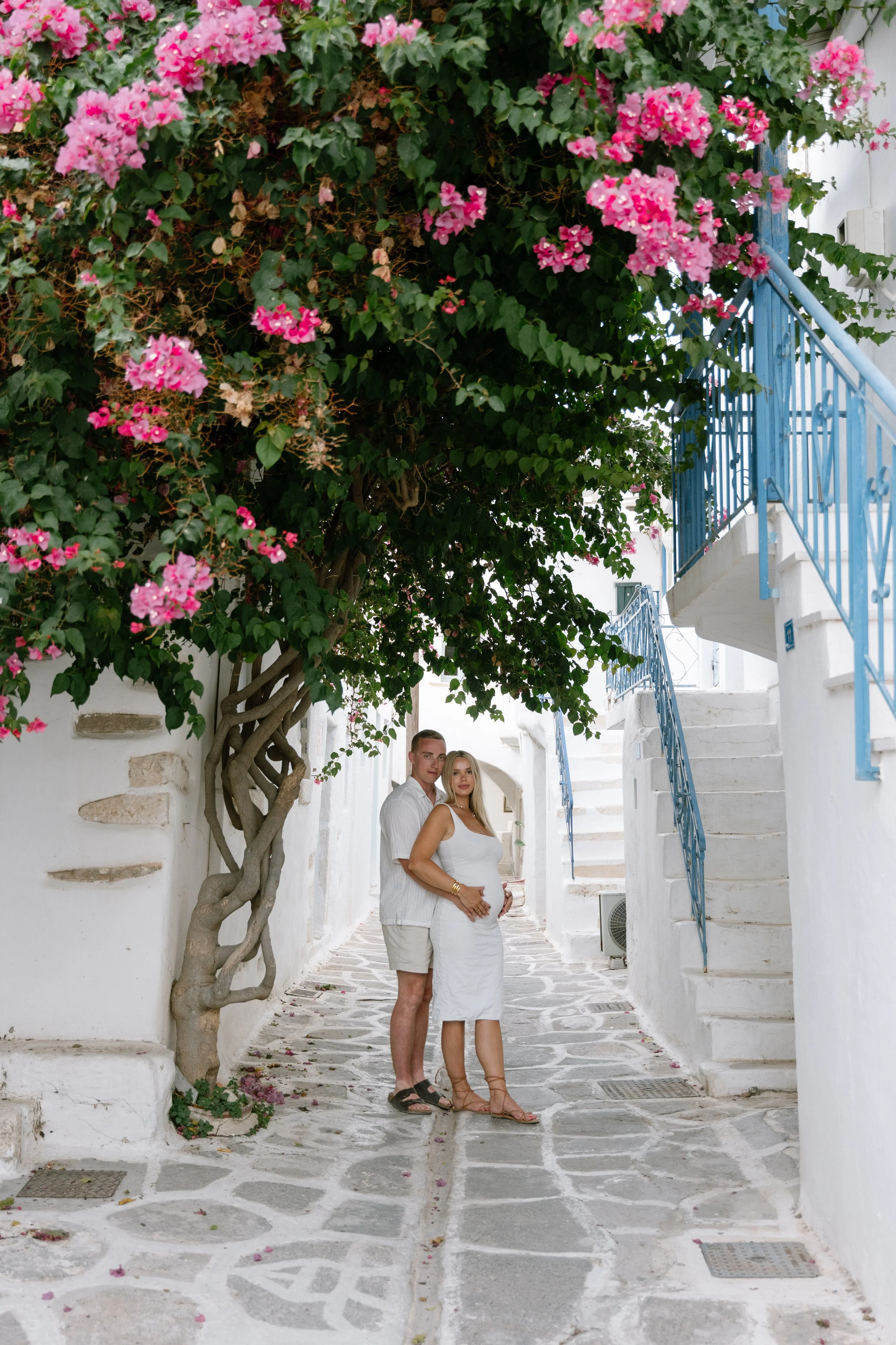 A couple standing on a cobblestone street underneath a large flowering green tree, with white buildings and blue railings in the background.