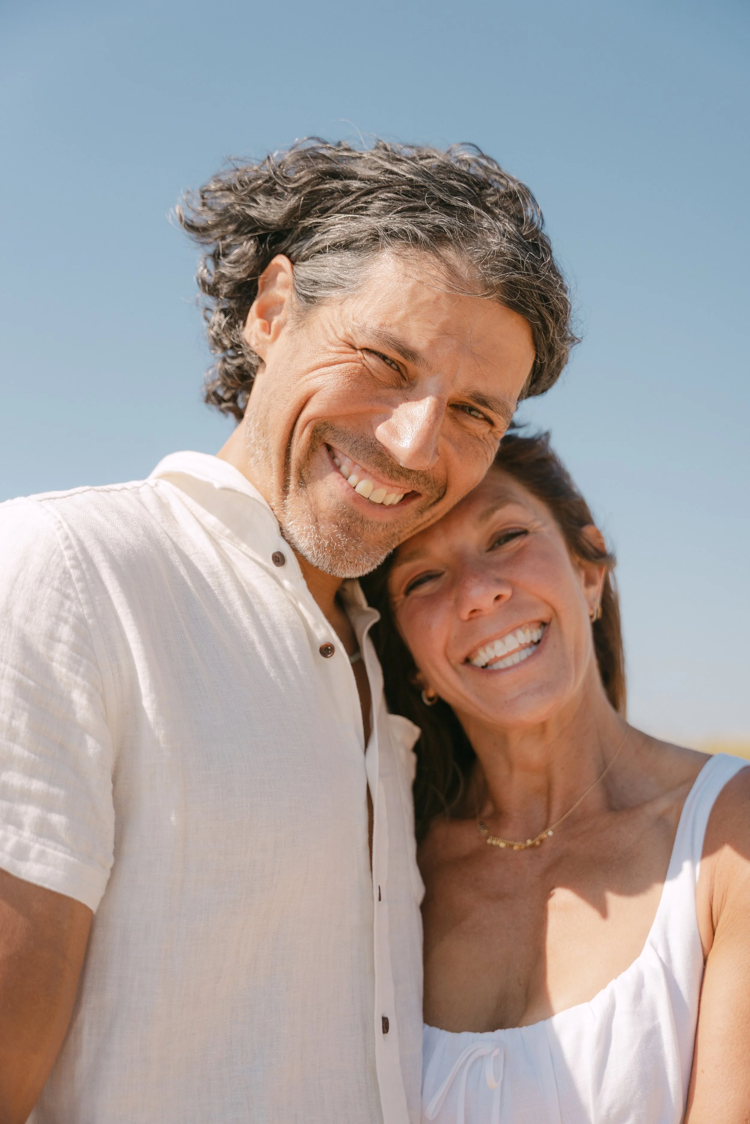 A smiling middle-aged couple taking a selfie outdoors against a clear blue sky, wearing light-colored clothing.