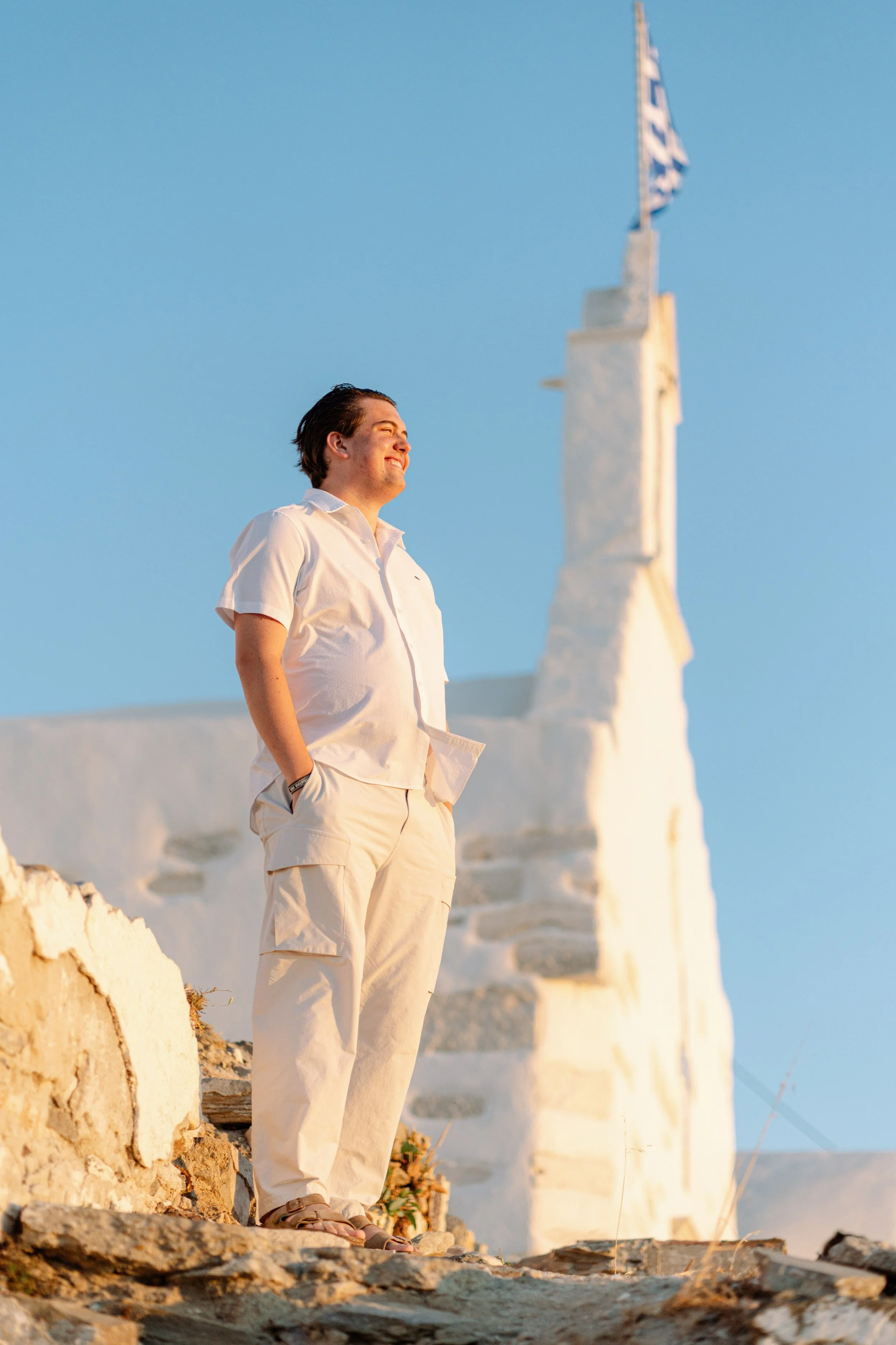 A man standing outdoors near a white building with stairs, under a blue sky, smiling as he looks into the distance.
