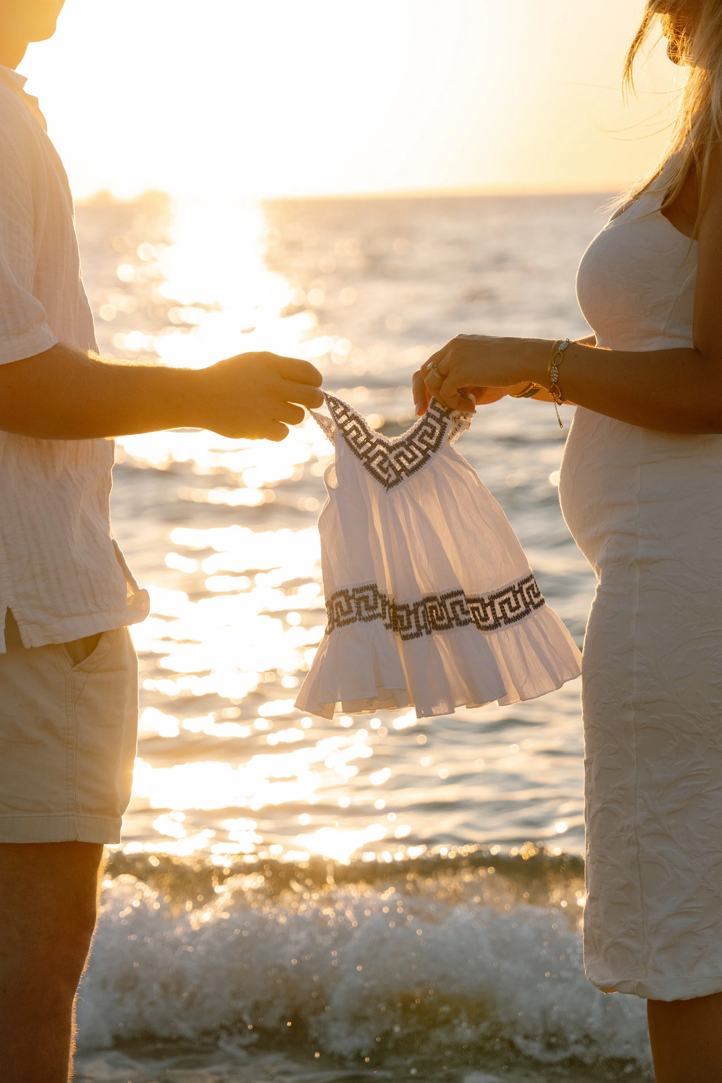 A couple standing in the ocean at sunset exchanging a white dress with black decorative patterns.