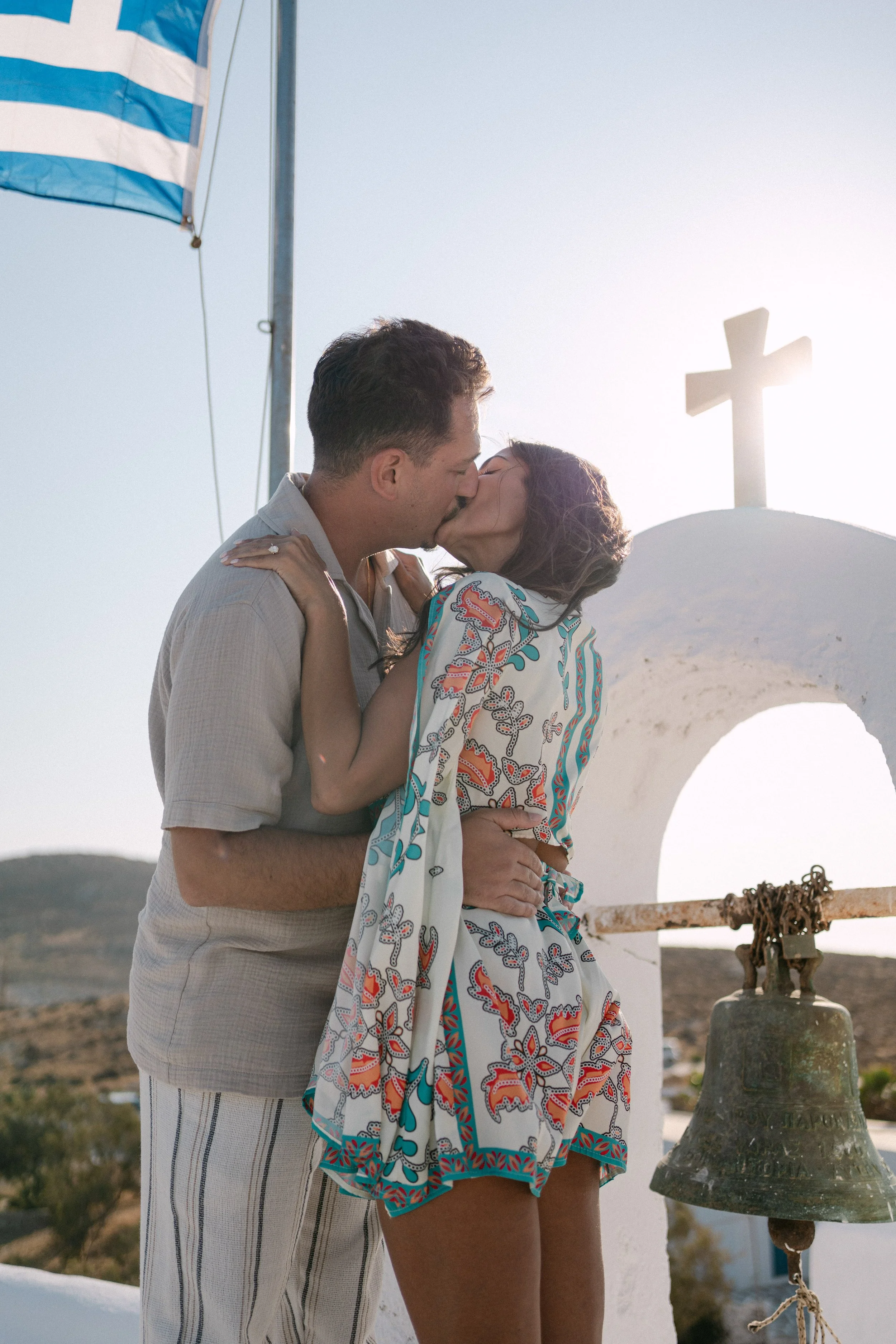 A couple kissing in front of a white chapel with a cross on top, with the man holding the woman around the waist and the woman with her arms on the man's shoulders. The wind blows her hair and her colorful dress. An American flag is visible in the ba