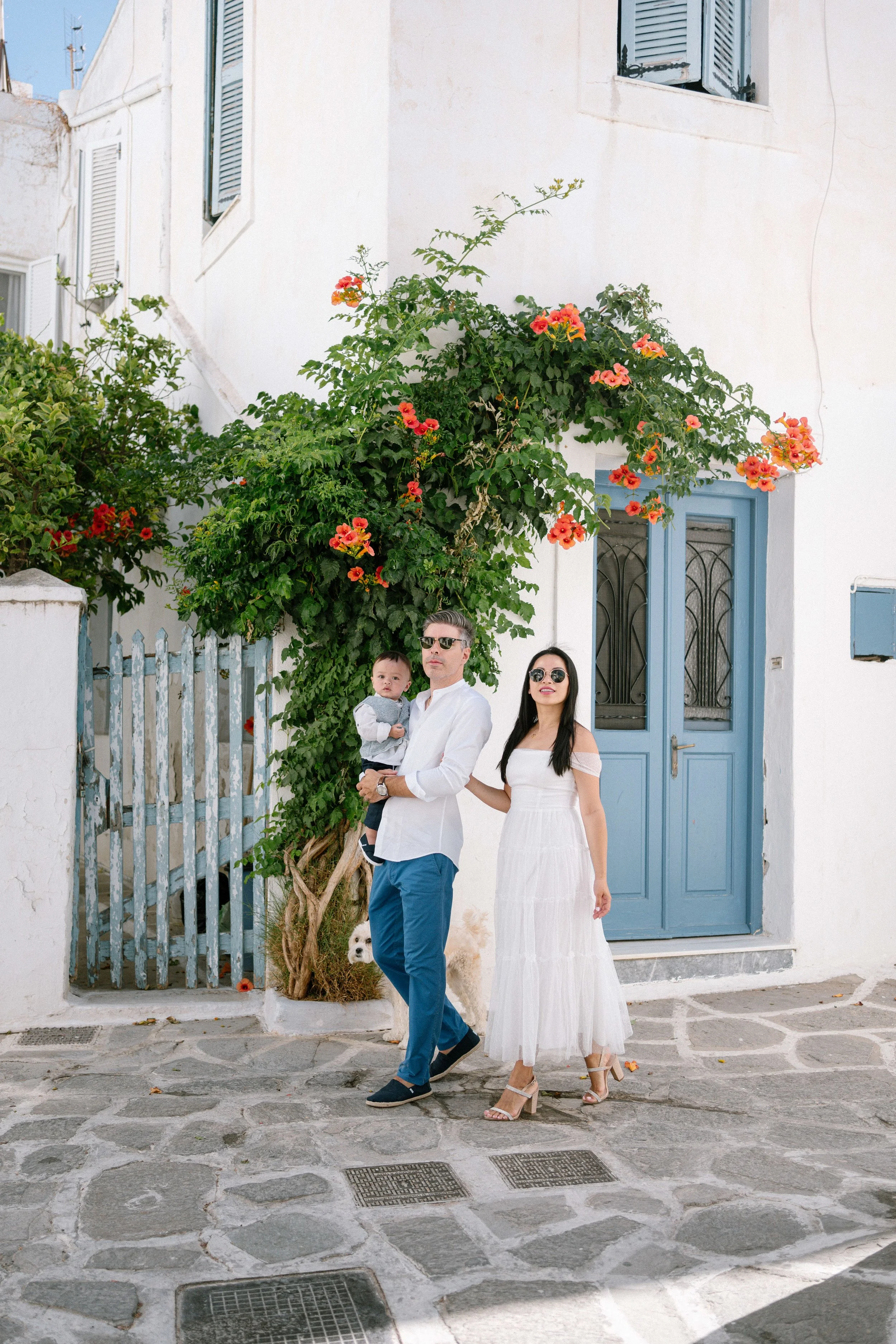 A couple and a young child standing on a cobblestone street in front of a white building with a blue door and window shutters. The man is holding the child, and they are all dressed in casual, stylish clothes with sunglasses. There is a flowering vin