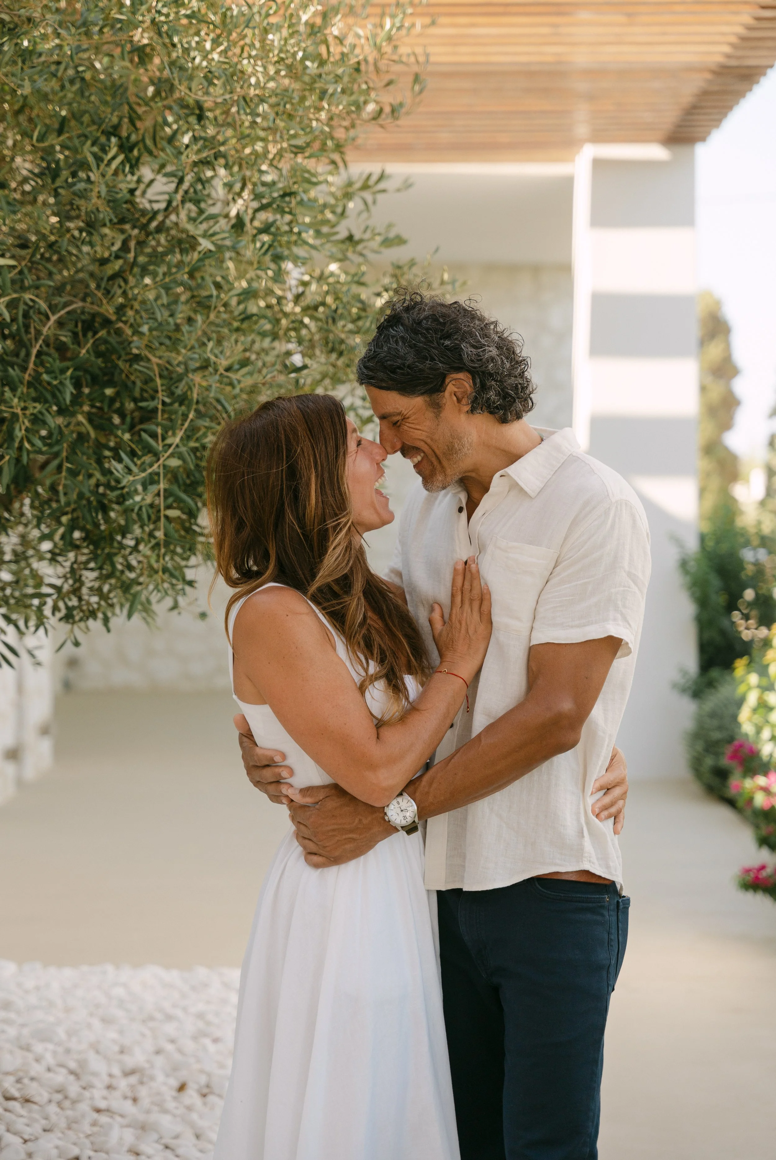 A happy couple is holding each other outdoors, touching noses and smiling, with greenery and modern architecture in the background.