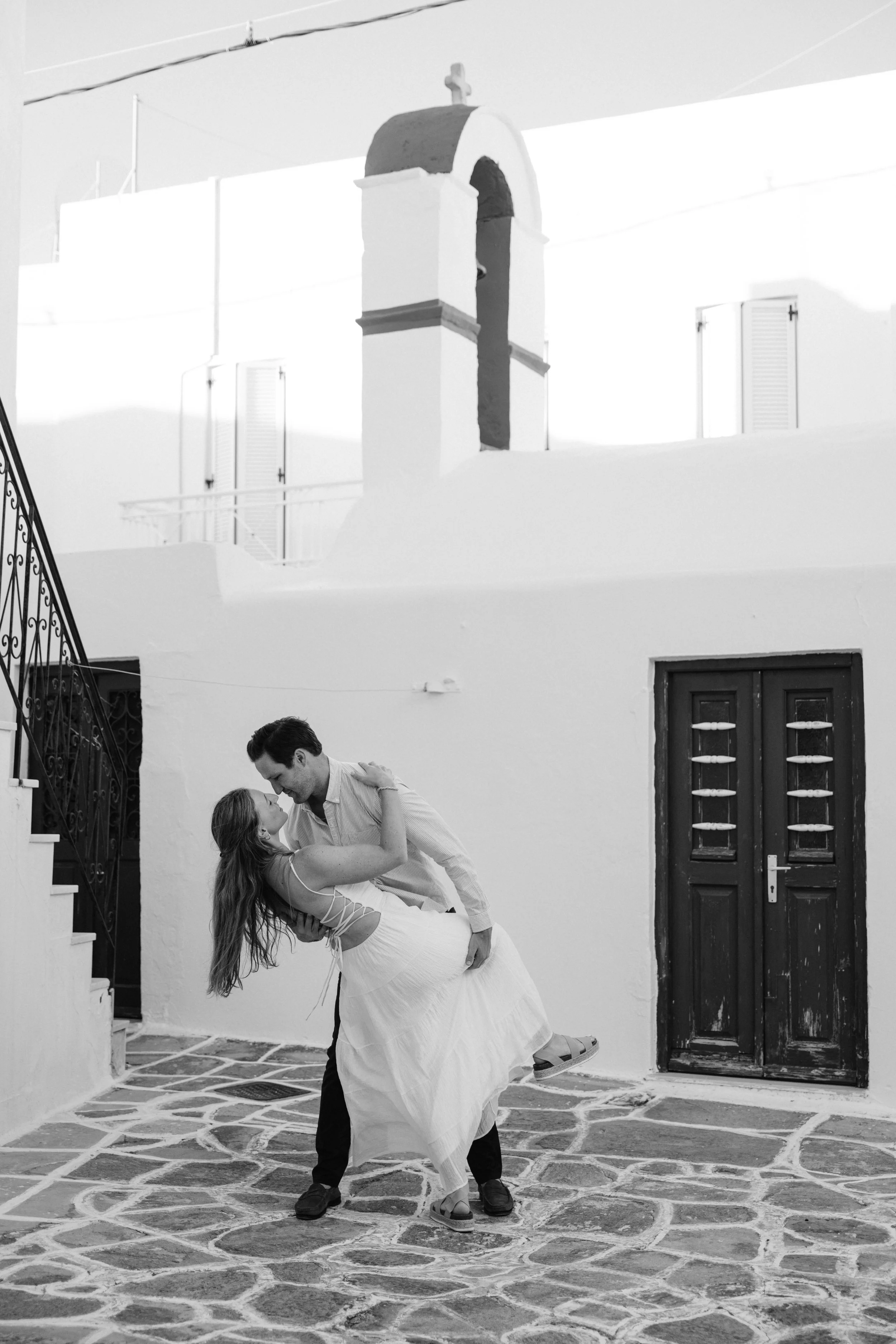 A man and woman dancing closely outside a white building with a dark door and spiral staircase, on a cobblestone street, in Greece.