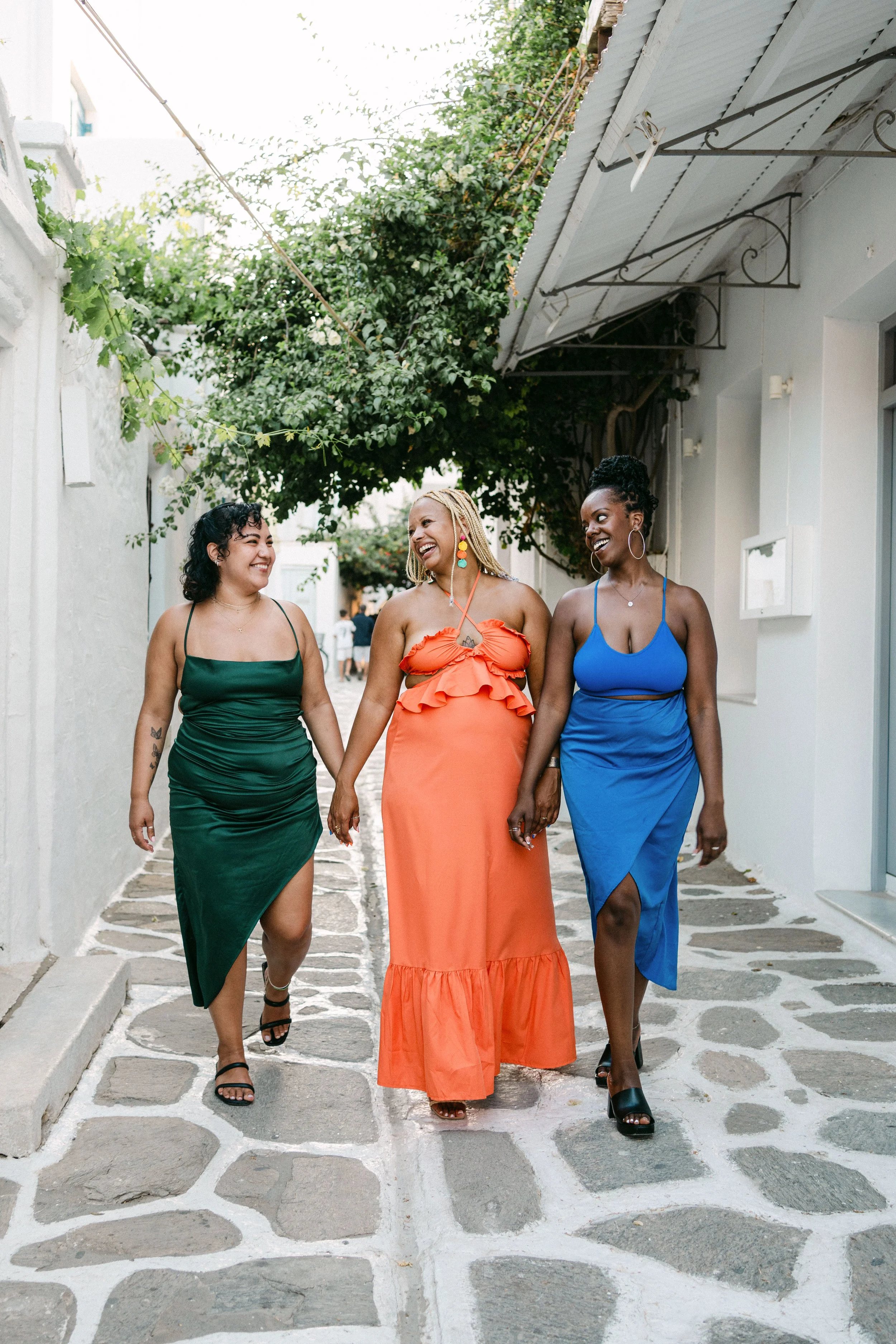 Three women walking together on a stone-paved street, smiling and holding hands, with white buildings and green foliage overhead, dressed in colorful summer dresses.