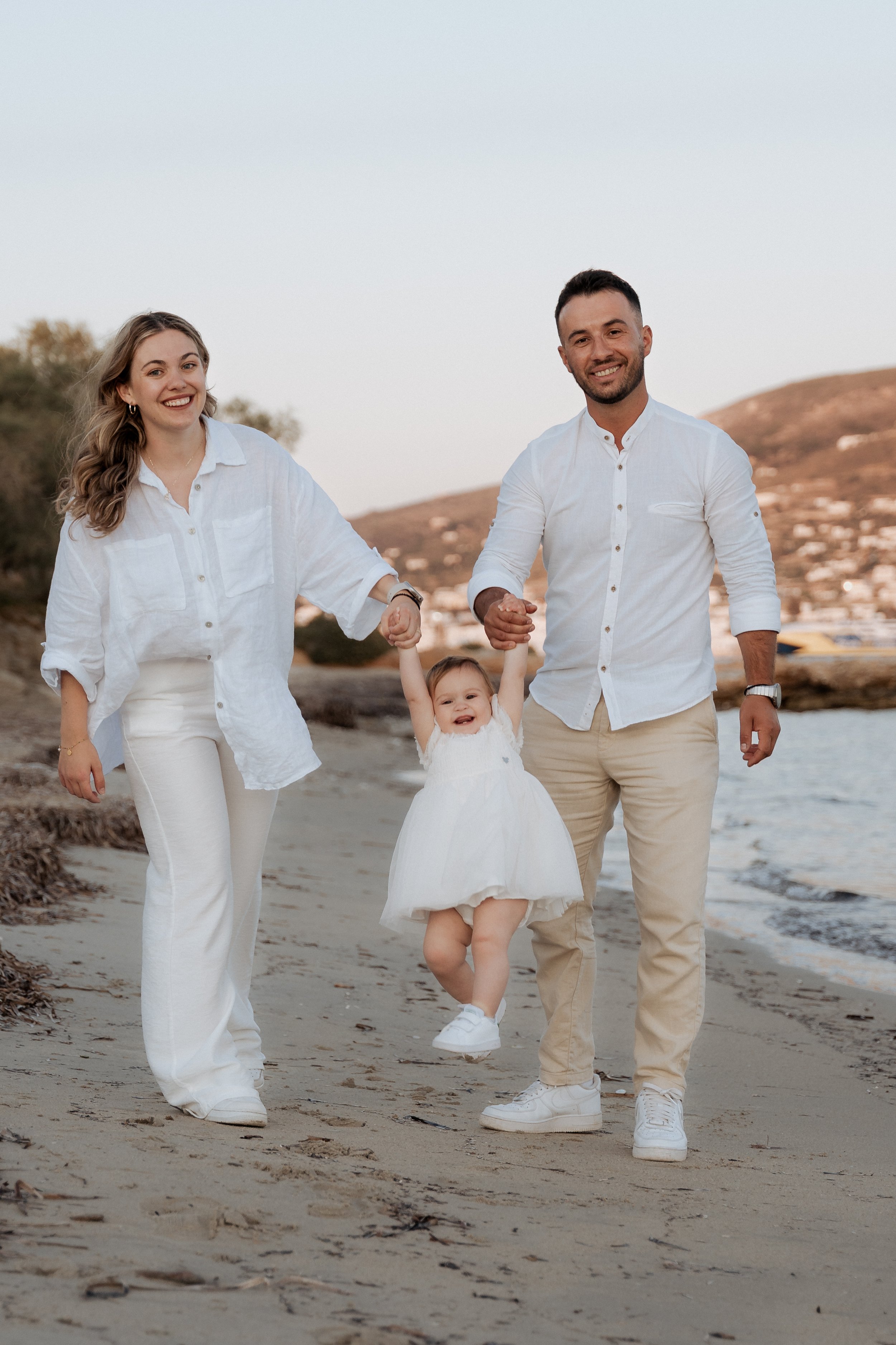 A happy family of three walking on the beach during sunset. The woman and man are holding the child's hands, swinging her in the air. They are all dressed in white with a scenic background of hills and water.