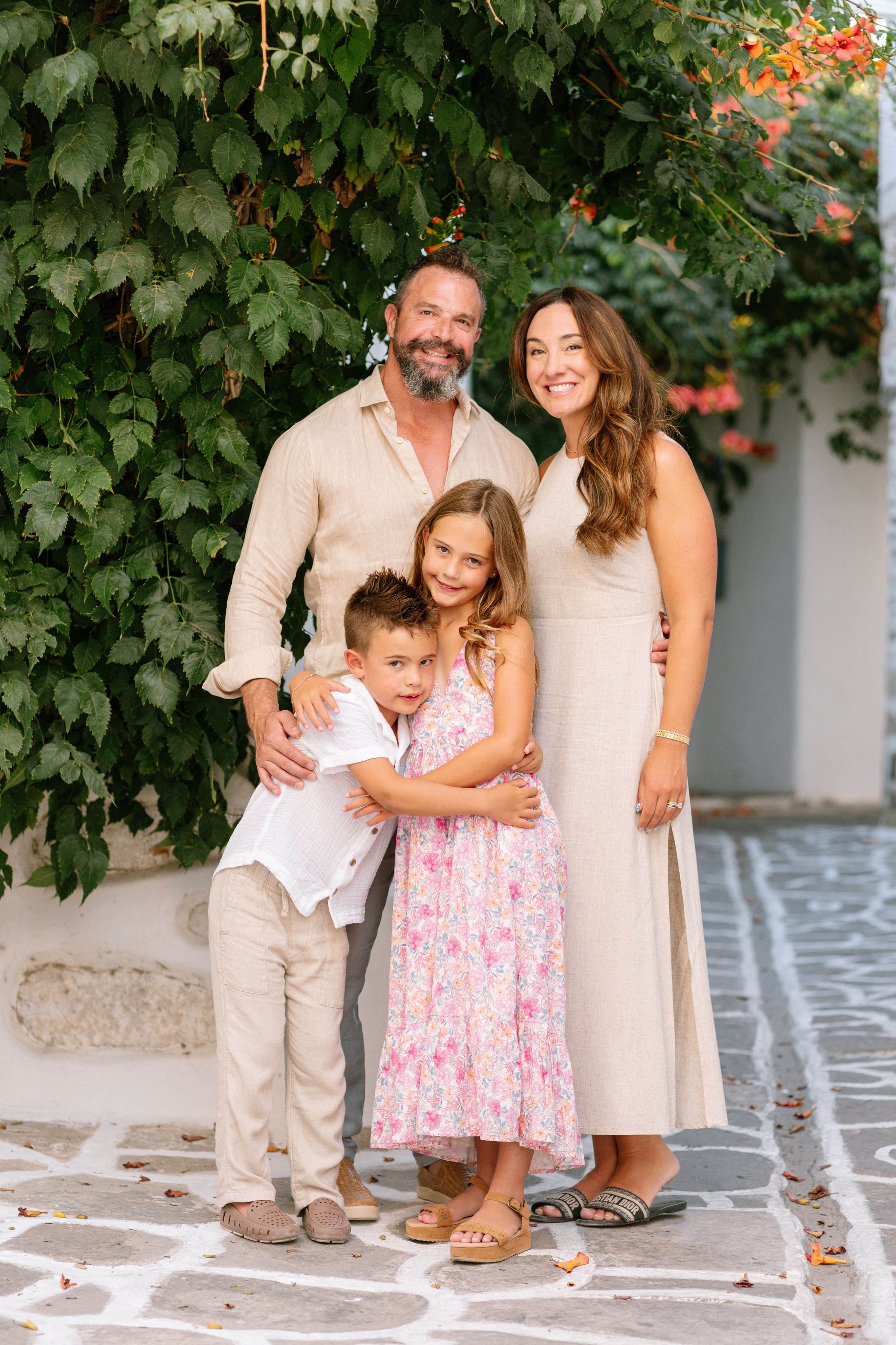 A smiling family of four standing together outdoors, with a lush green leafy and flowering background.