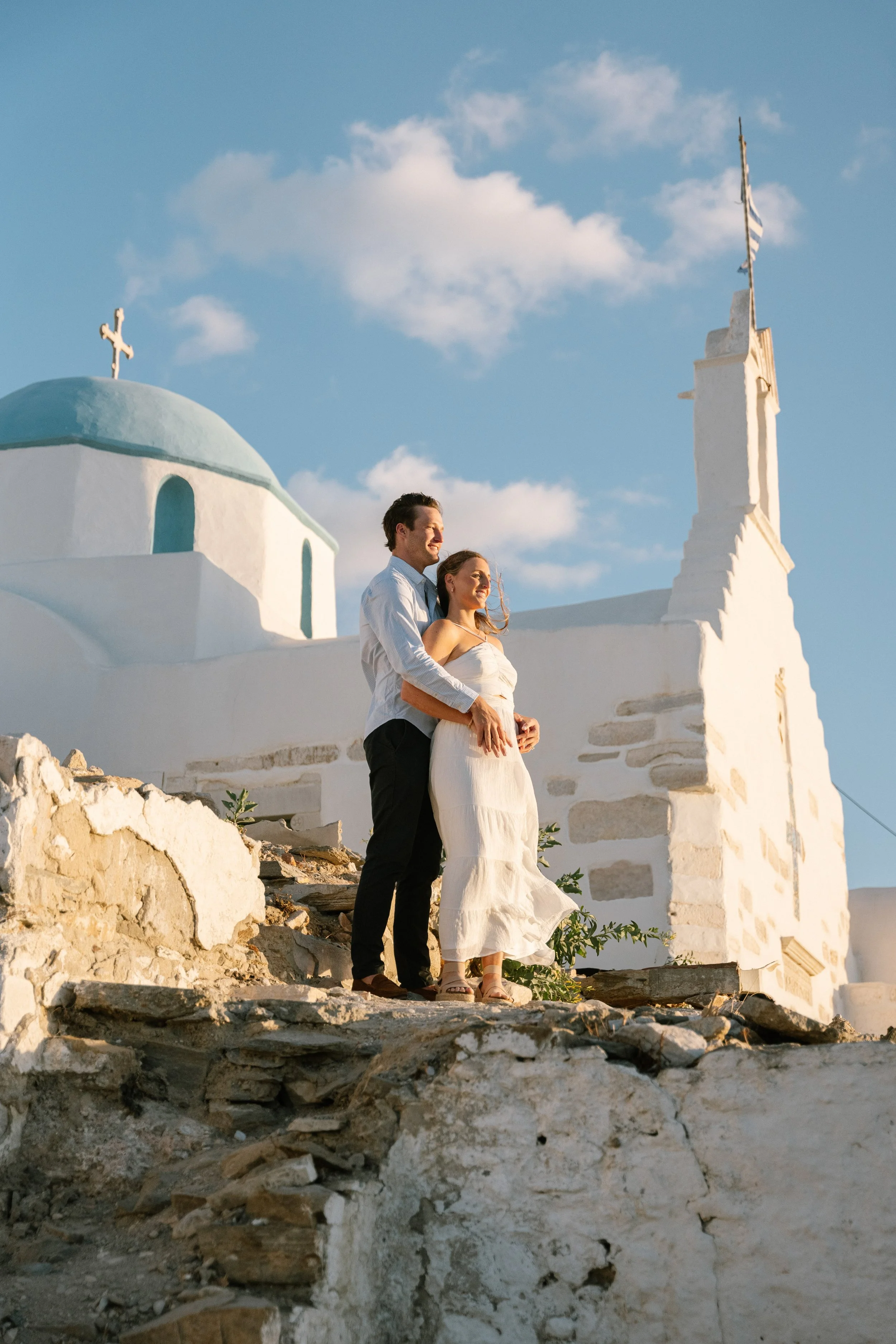 A couple stands on rocky terrain near a white church with a blue dome in the background, under a blue sky with clouds, during sunset.