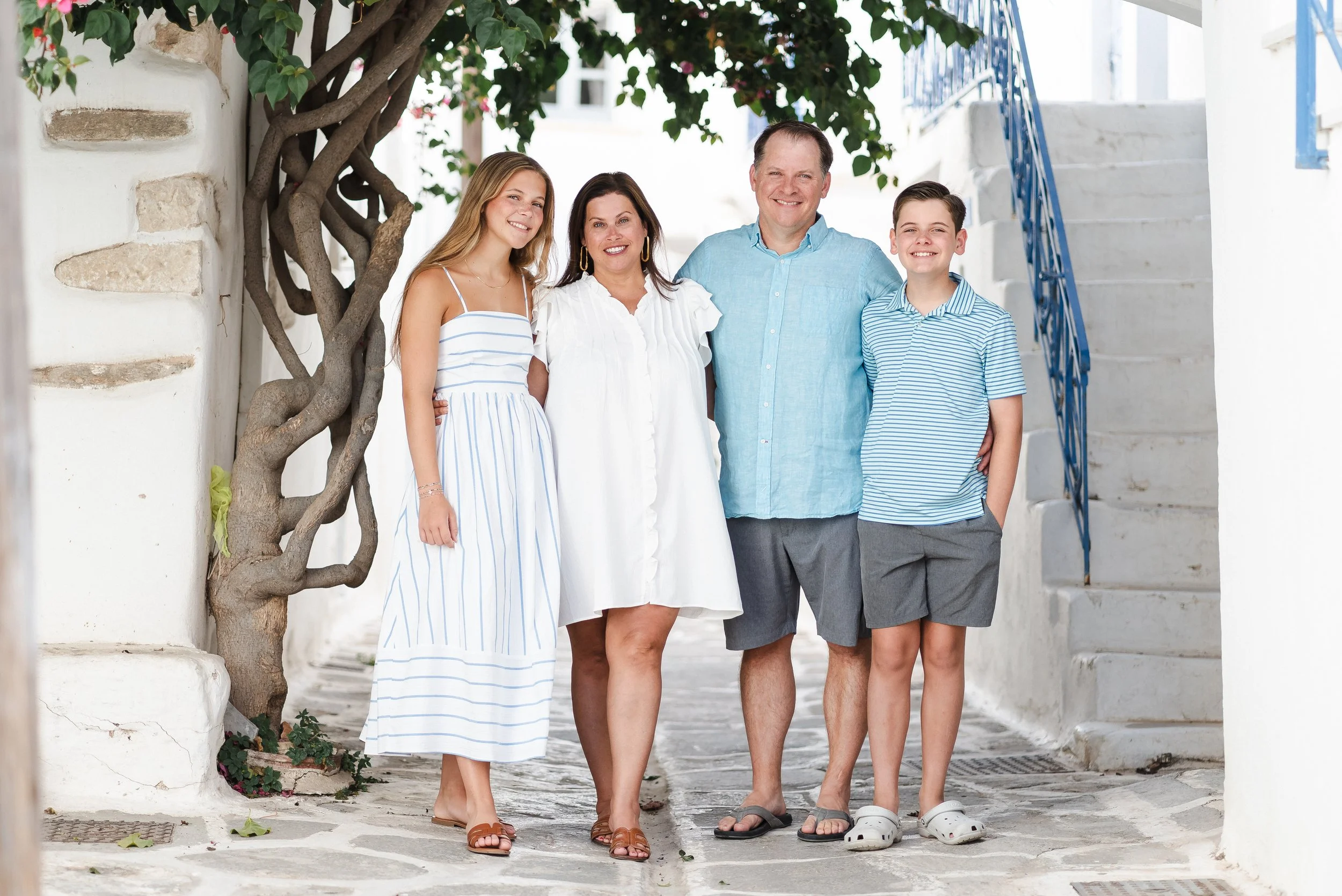 A family of five standing close together outside on a cobblestone street, smiling for the camera, with white buildings and a blue staircase in the background.