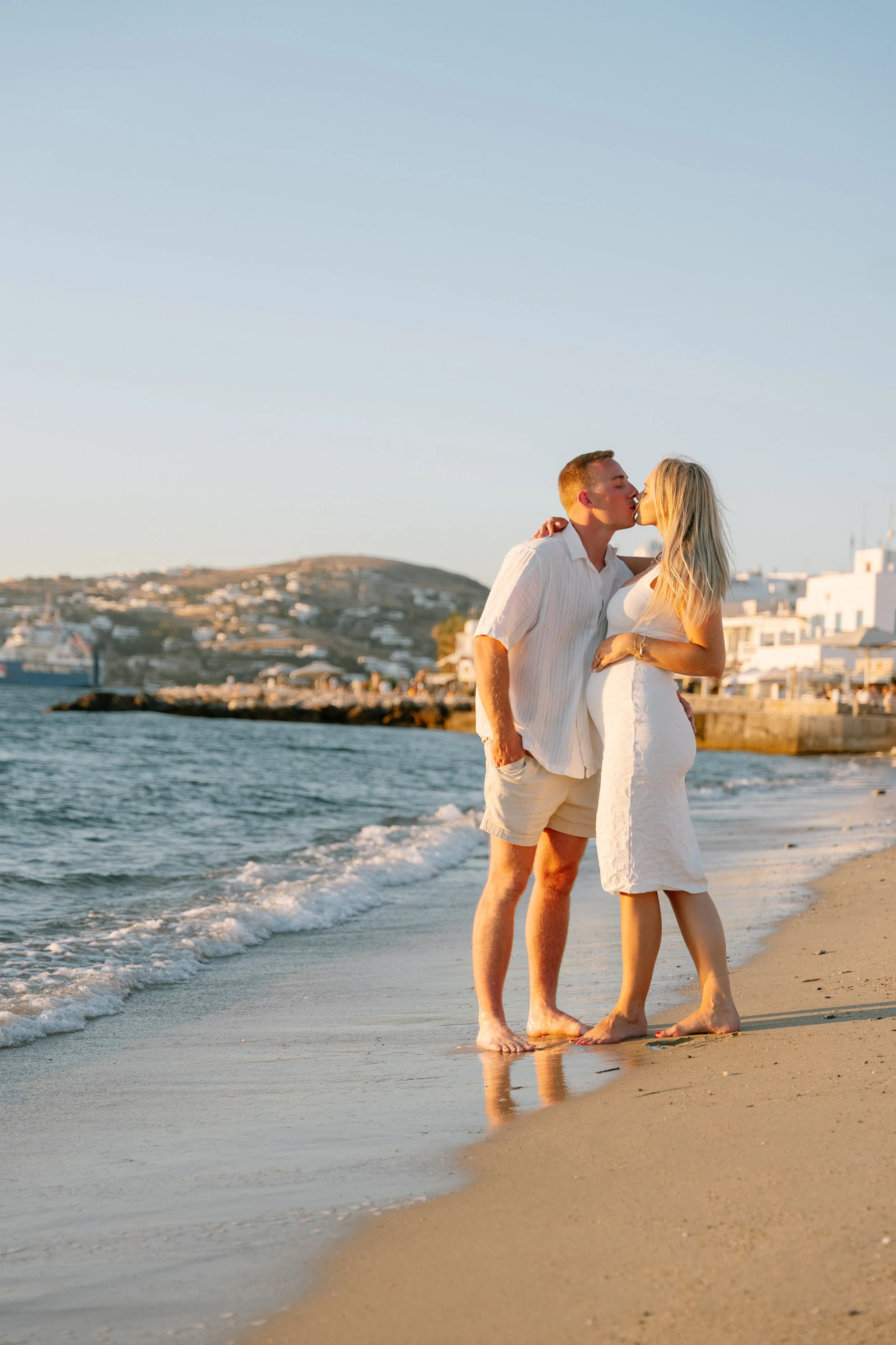 A couple sharing a kiss on the beach during sunset, with a seaside town and hills in the background.