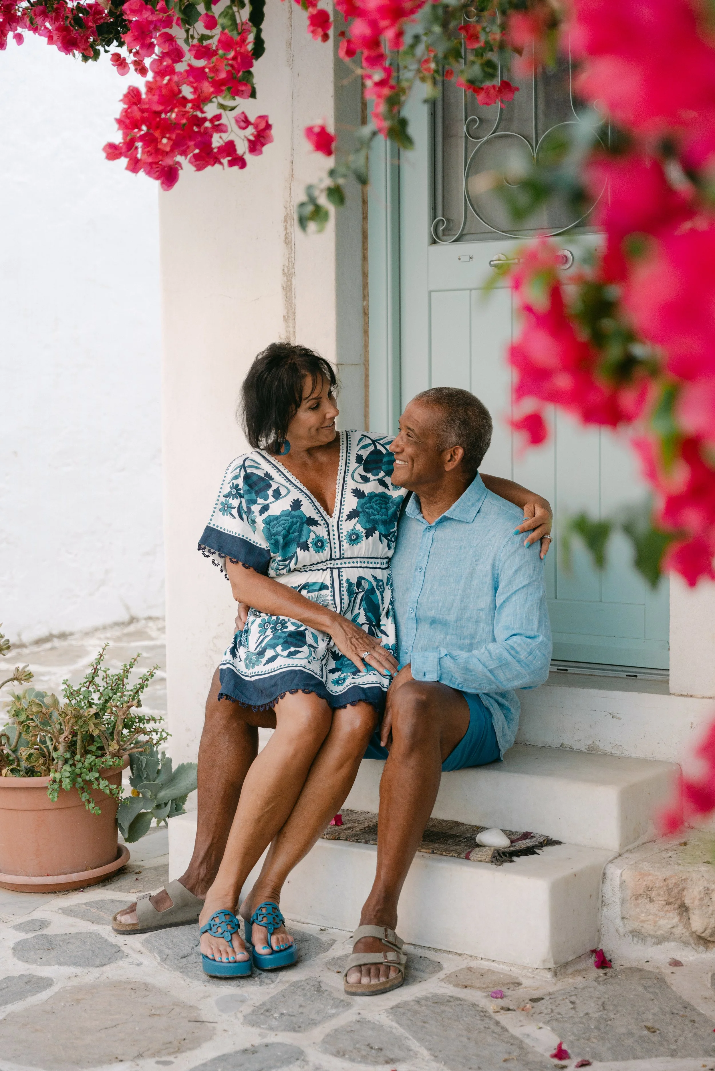 A couple sitting on the steps of a house, smiling and embracing, with pink flowers hanging and blooming around them.