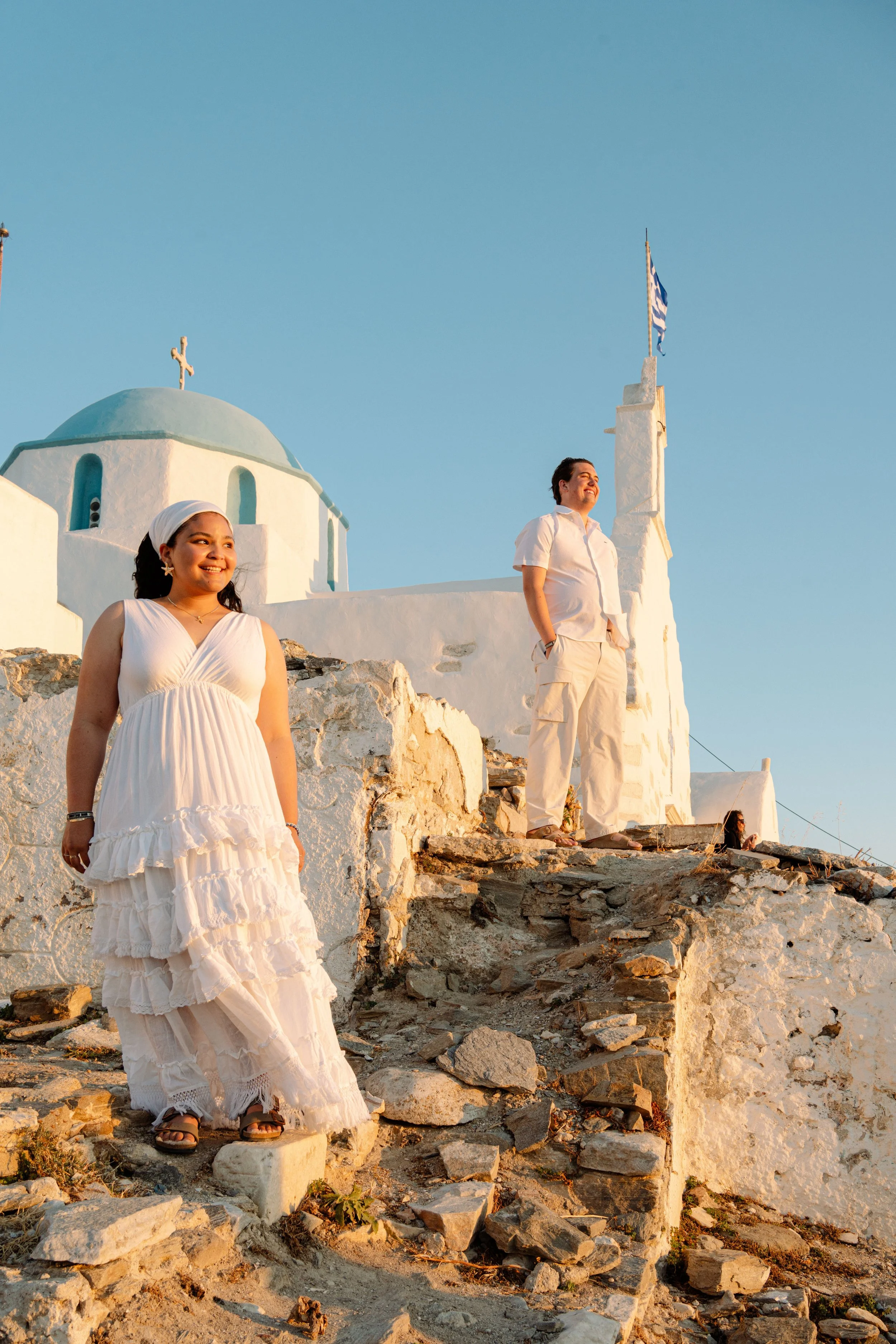 Two women standing on rocky steps in front of white buildings with blue domes and a cross, during sunset in Greece.