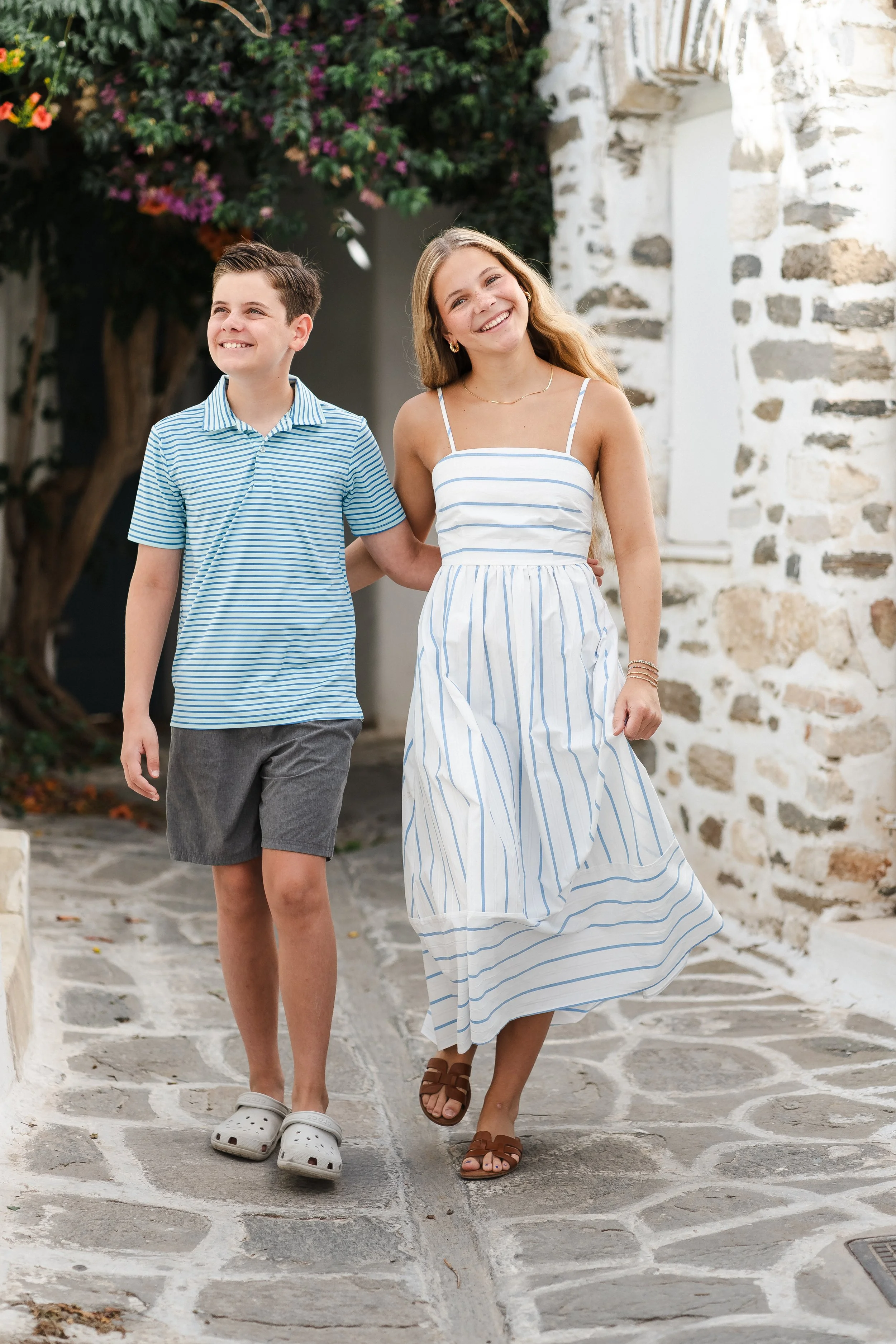 A woman and a boy walking together outdoors on a stone pathway, smiling, with a stone building and flowering shrub in the background.
