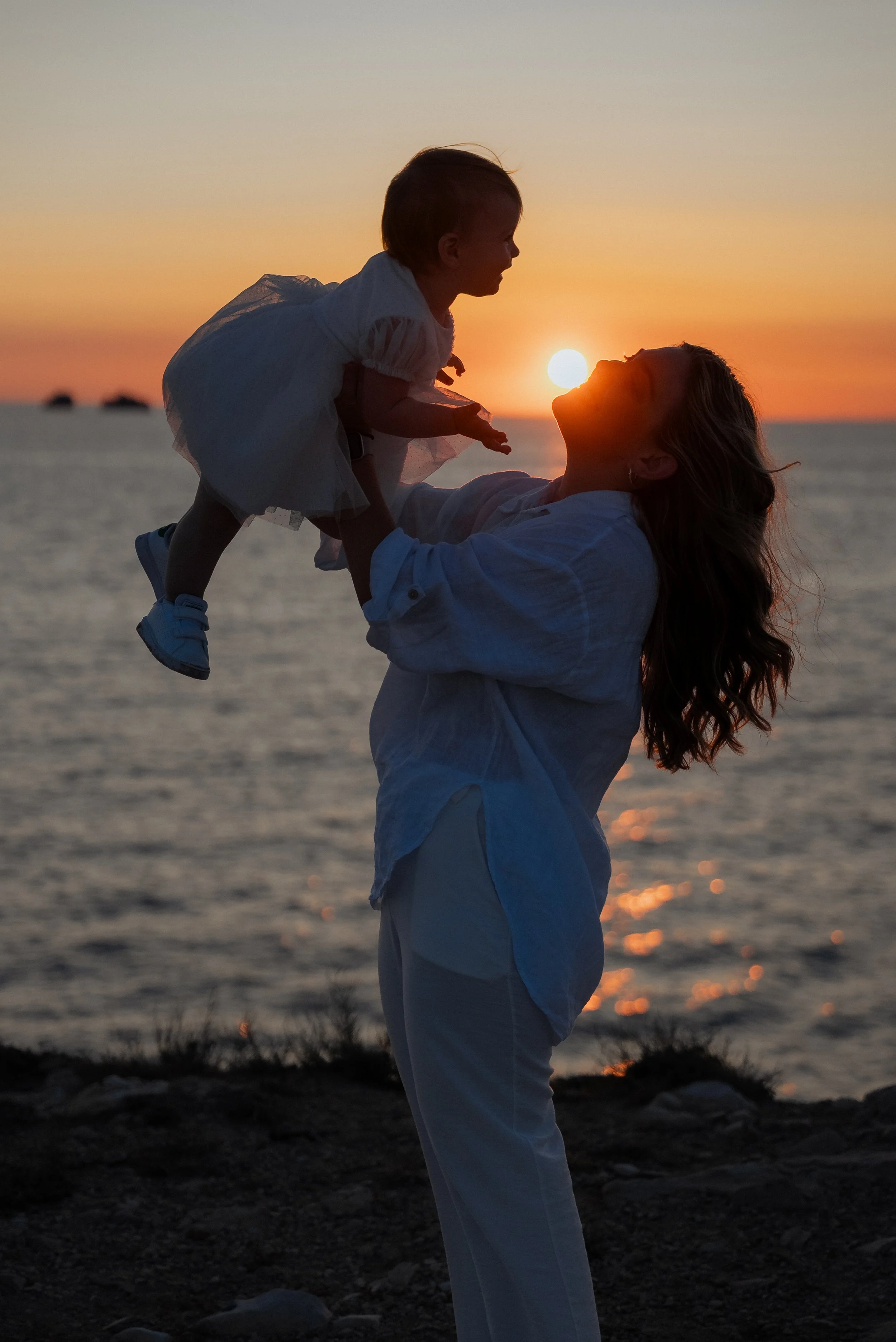 A woman lifting a child at sunset on the beach, with the sun setting over the ocean in the background.