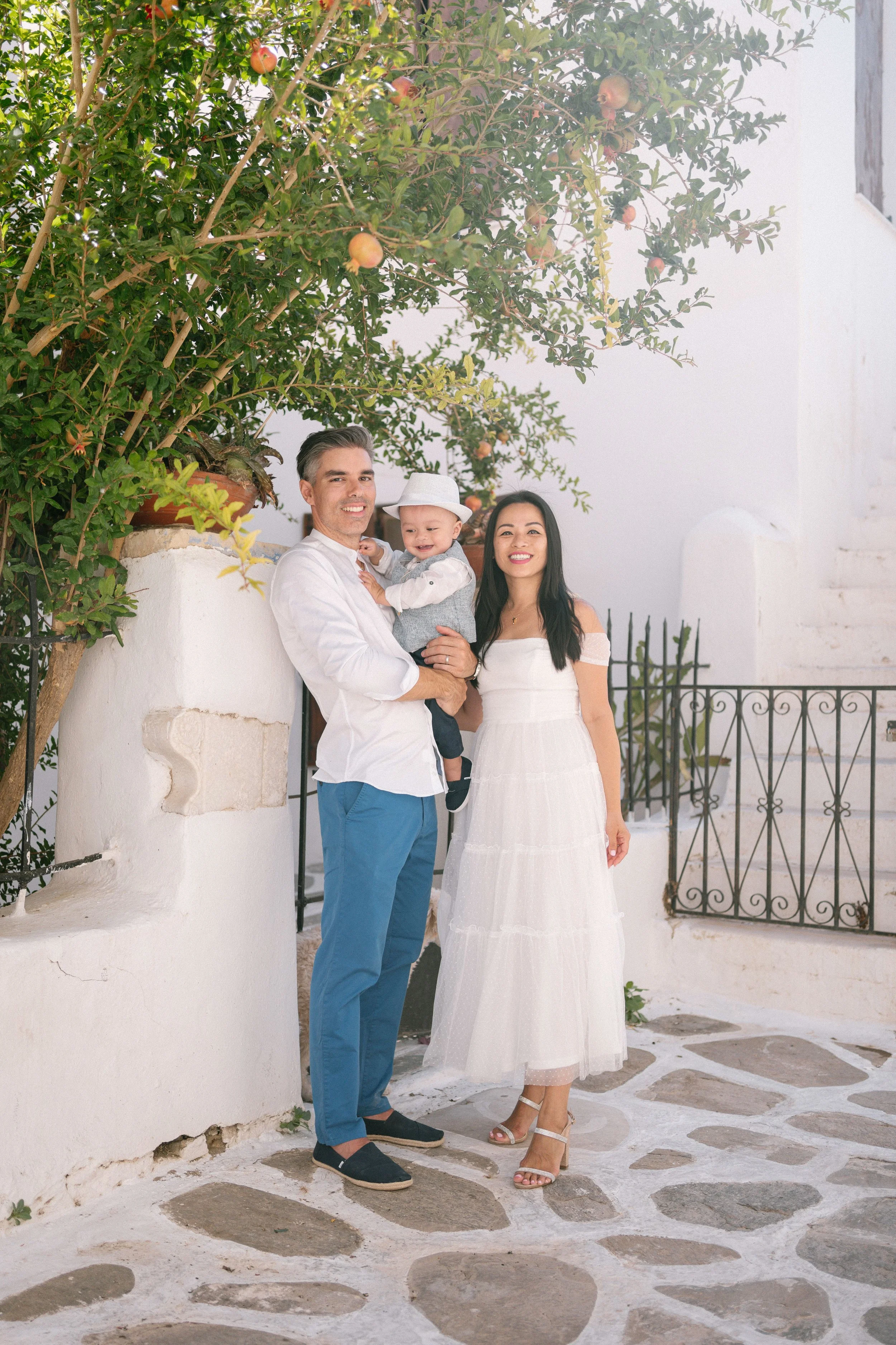A family of three, including a man, woman, and a young boy, standing on a stone-paved area in front of a white wall and railing. The man holds the boy, who is wearing a hat, and all are smiling. There is a large pomegranate tree with green leaves and