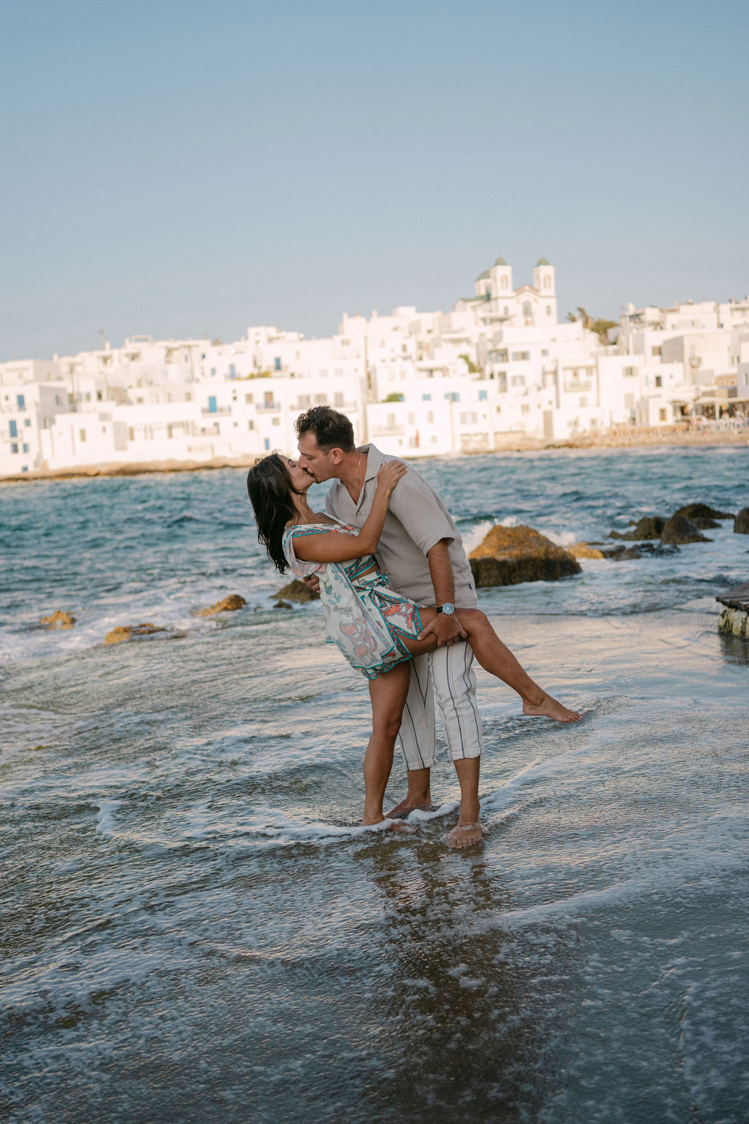 A couple sharing a kiss on the beach at sunset, with white buildings and a church on a hill in the background.