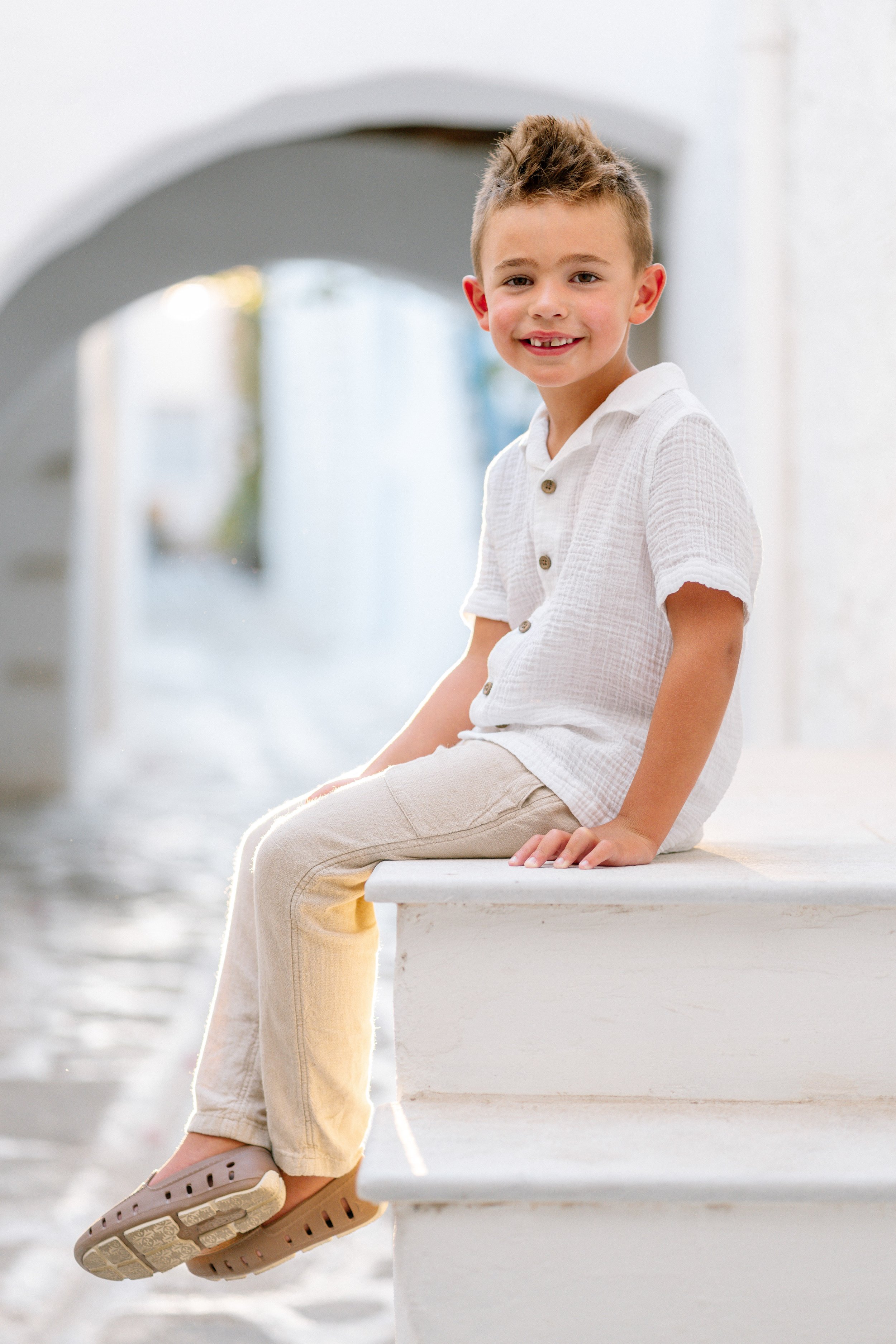 Young boy sitting on a white ledge near a canal or waterway, smiling at the camera, wearing a white short-sleeve button-up shirt and beige pants.