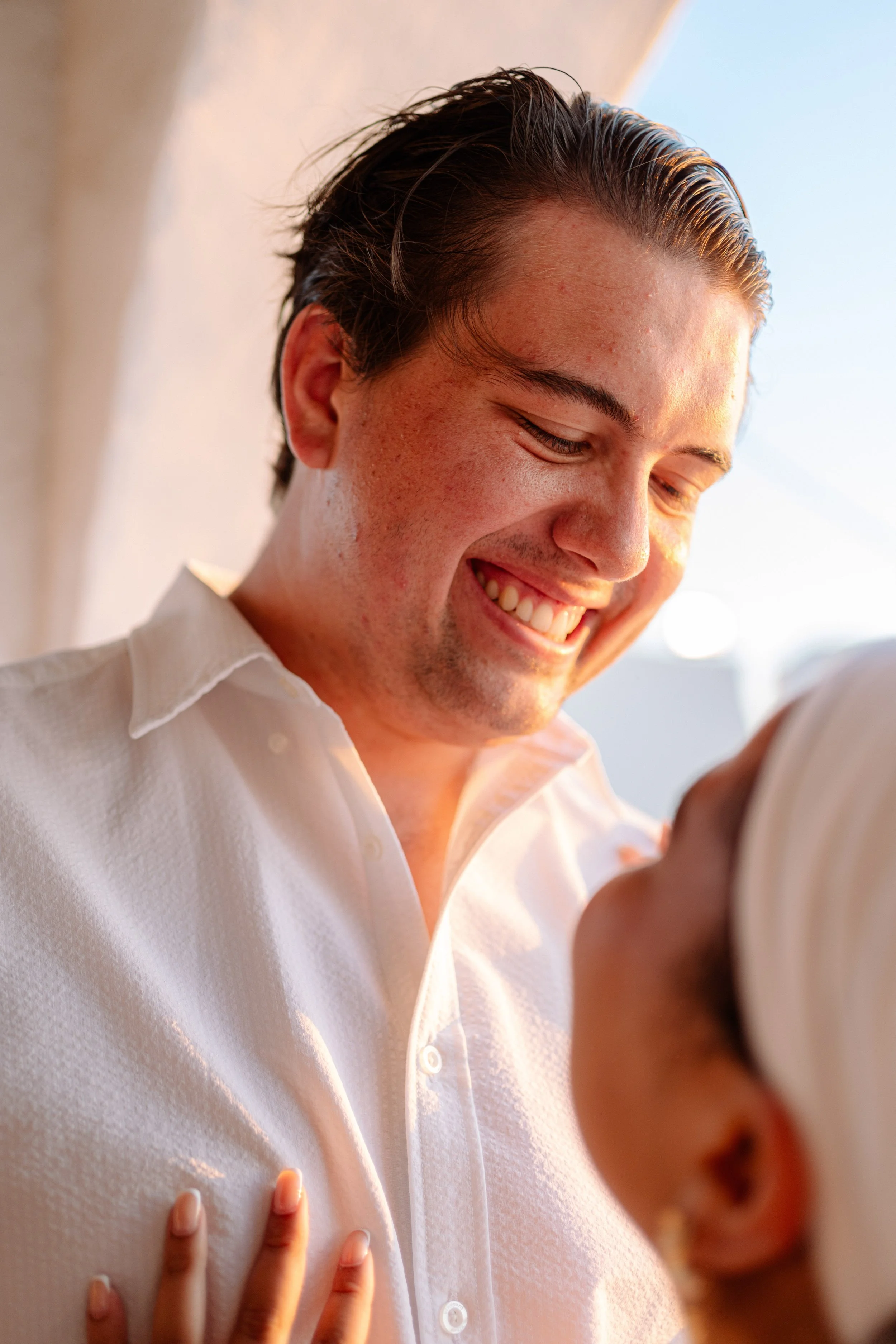 A young man with dark, wet hair smiling as he looks at a camera, wearing a white shirt, with a hand touching his chest.