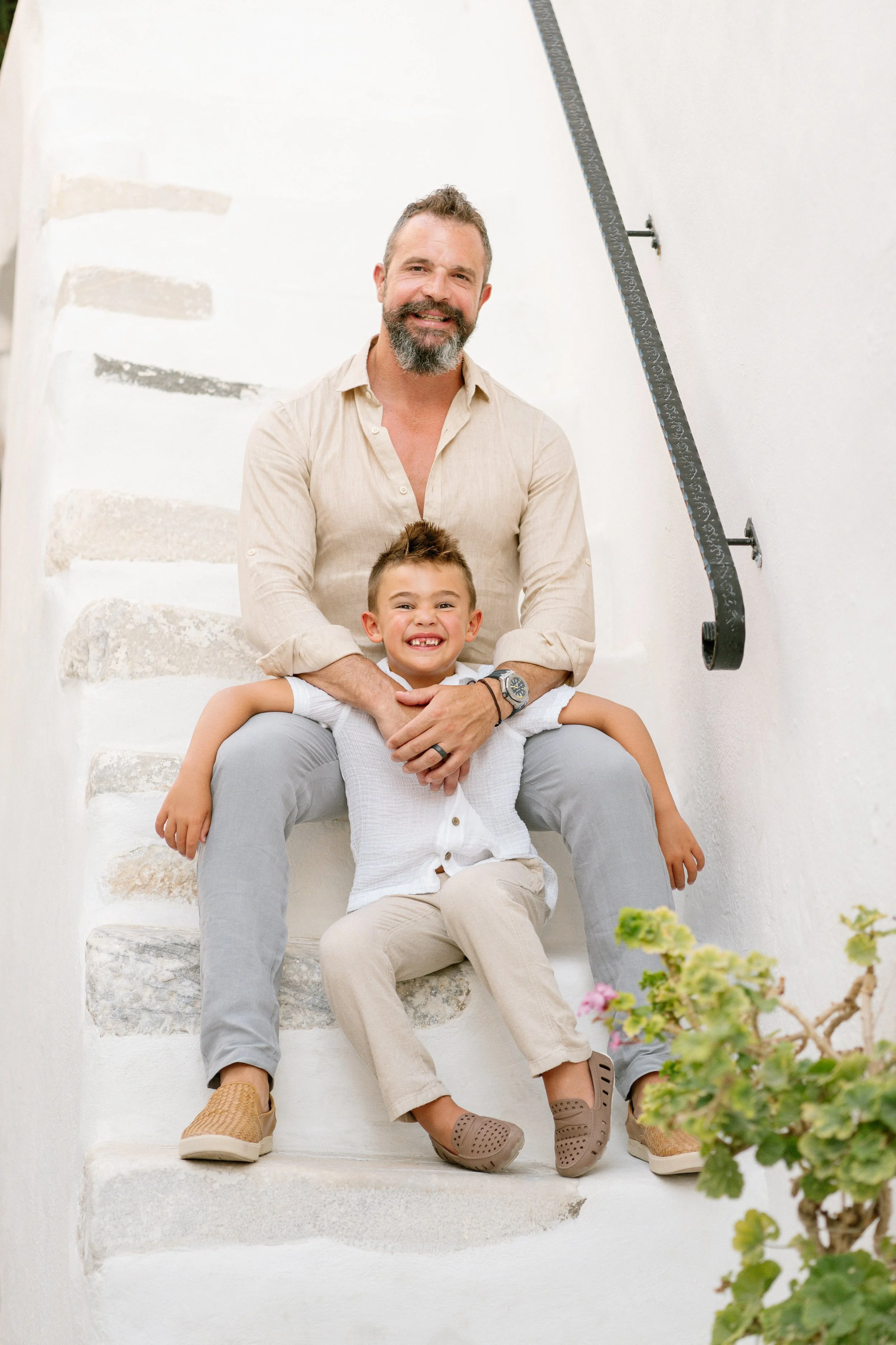 A smiling man and boy sitting on white stone stairs, with the man holding the boy from behind.