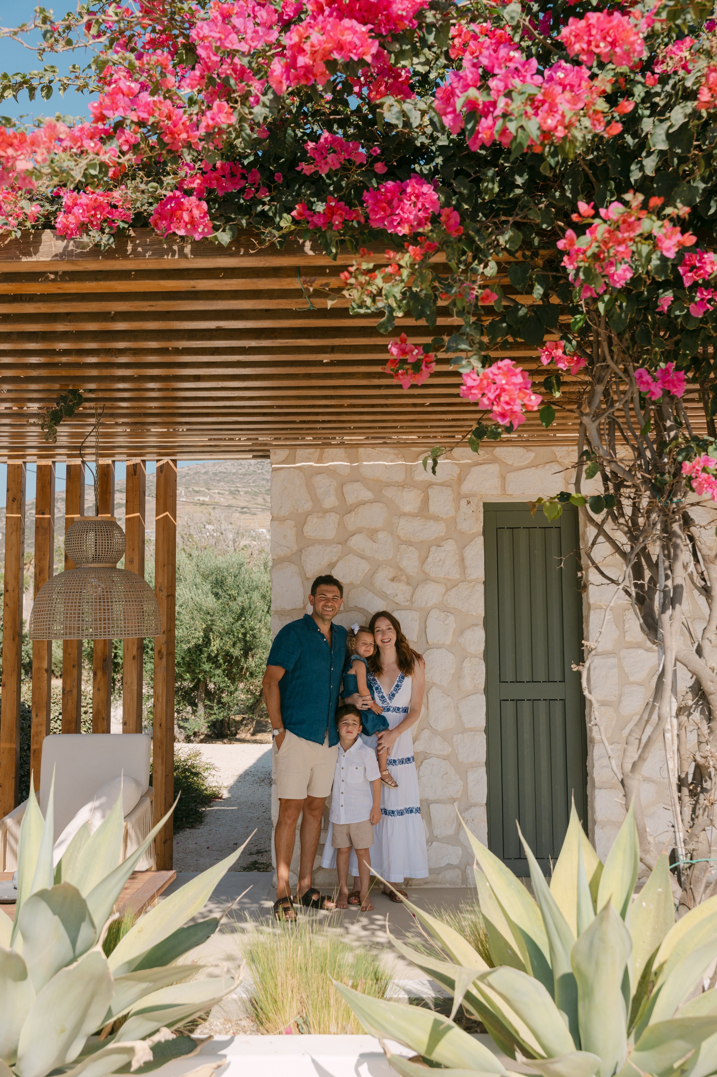 A family of four standing outside a stone building with greenery and pink flowering bushes overhead.