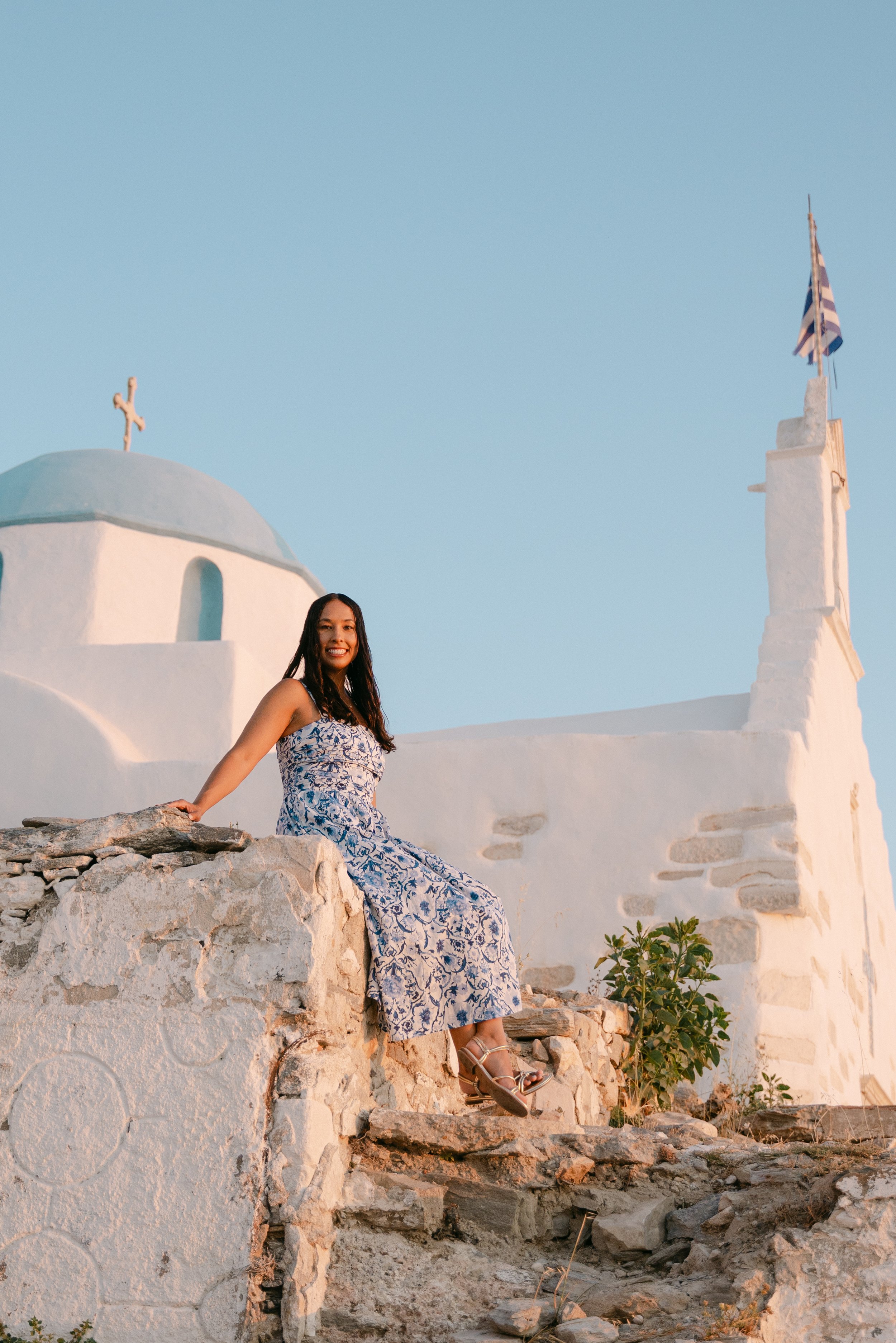 A woman in a blue-and-white dress sitting on a stone wall in front of white buildings, with a cross and a Greek flag on a building in the background, during sunset.