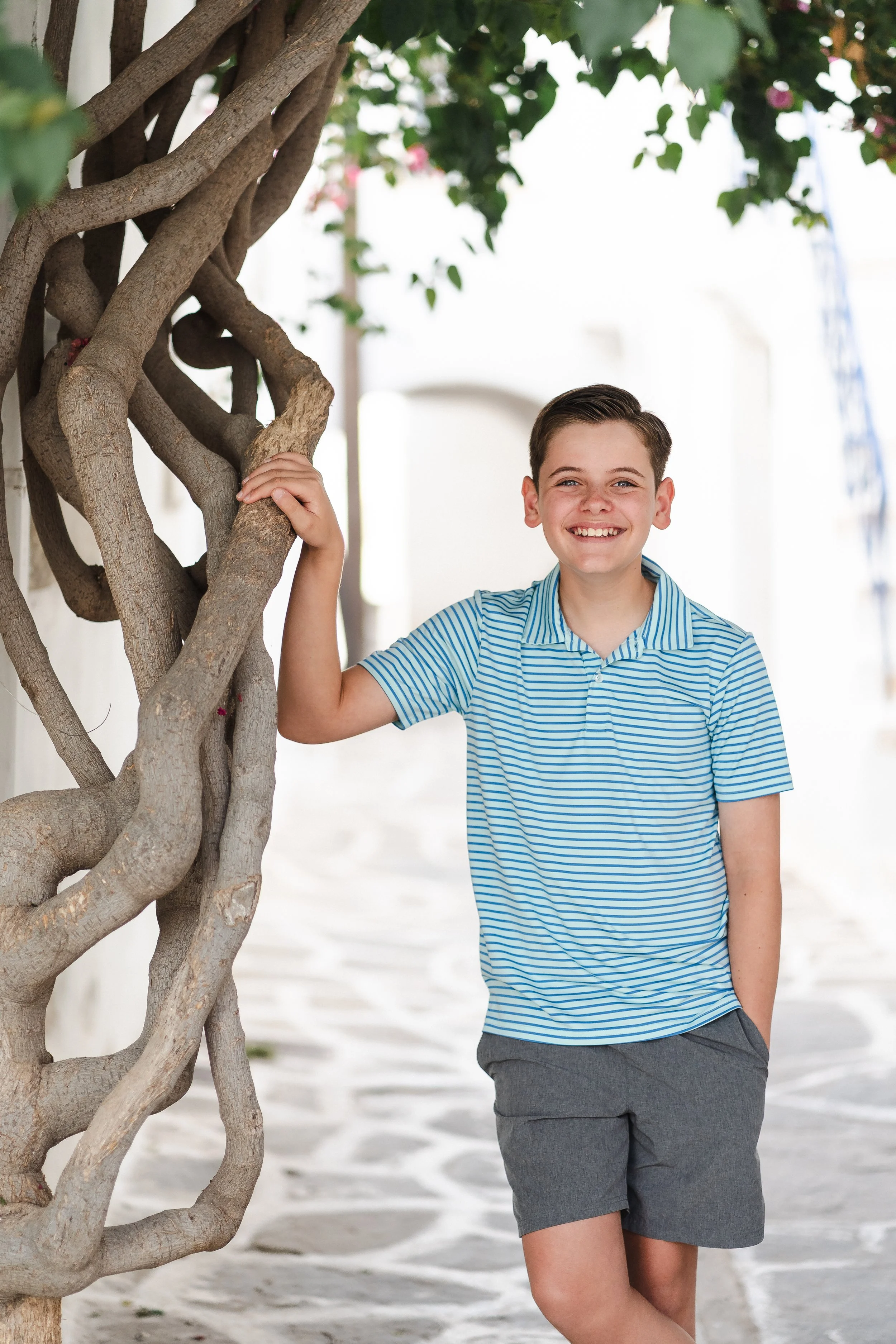A smiling young boy in a blue and white striped polo shirt and gray shorts, standing outdoors next to a twisting tree with green leaves.