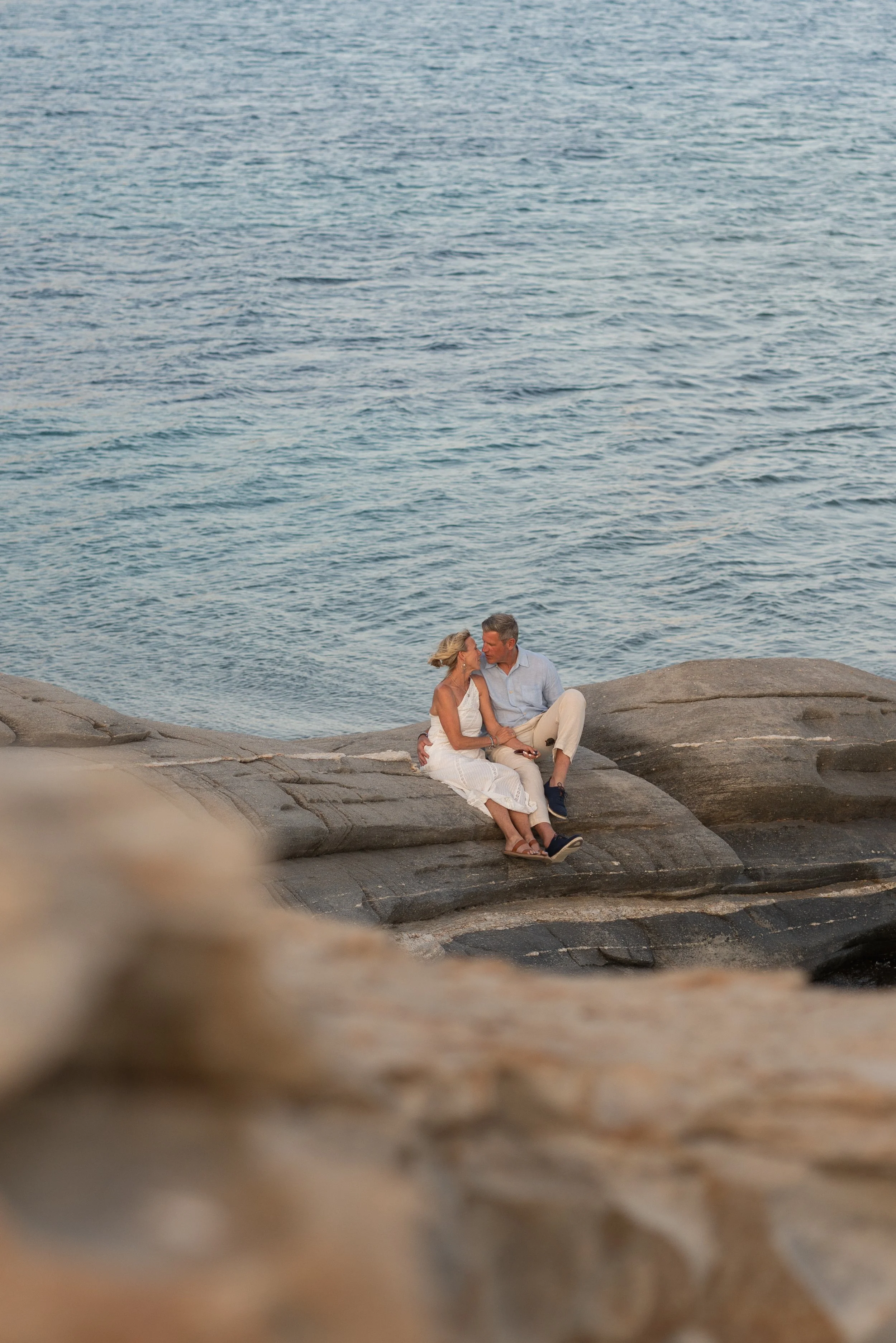 A couple sitting closely together on a rocky shoreline near the water, sharing an intimate moment at sunset.