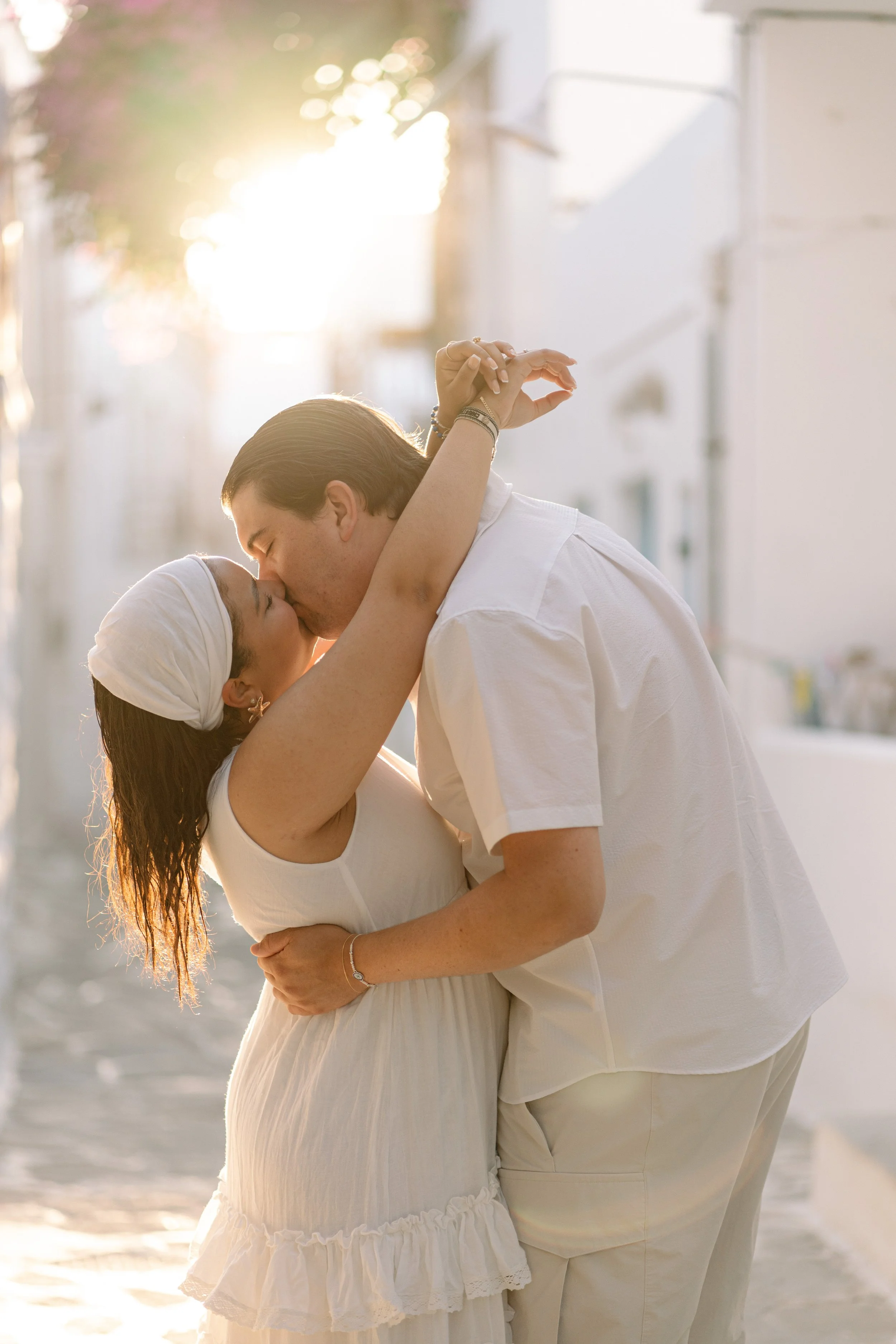 A couple kissing on a sunlit street during sunset, dressed in white, with the woman wearing a headscarf and the man leaning in to kiss her.