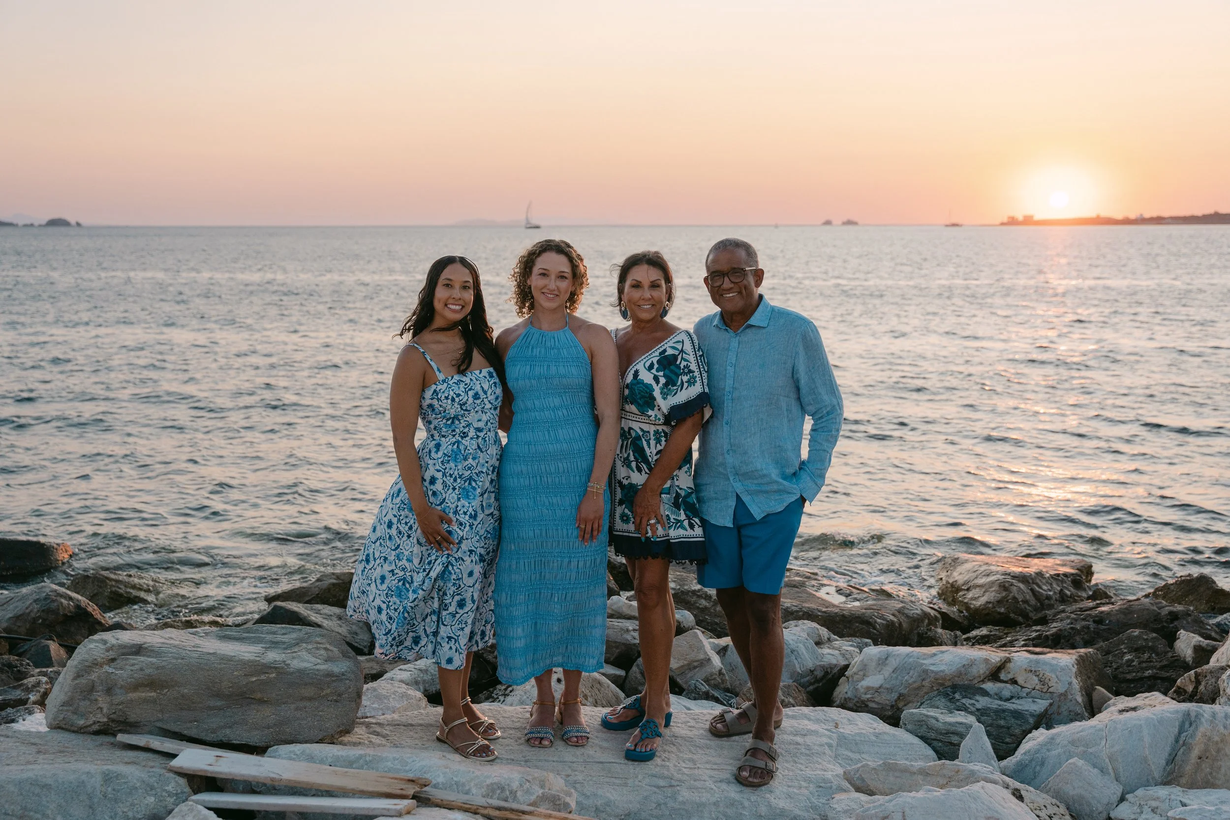 Group of four people standing on rocks by the ocean at sunset, smiling, dressed in casual summer clothing.